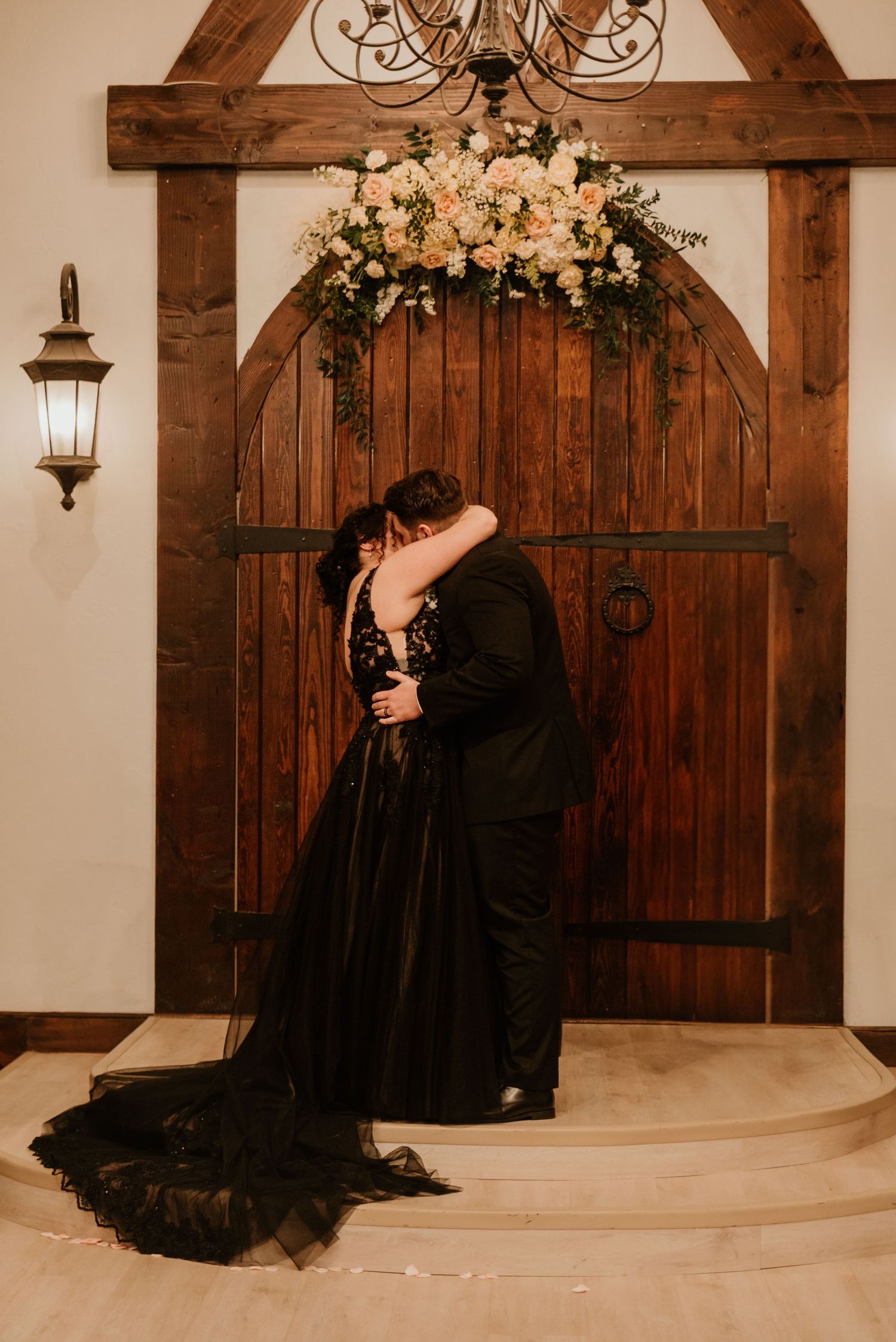 Bride and groom share first kiss beneath floral arch over rustic arched wooden doors after ceremony.