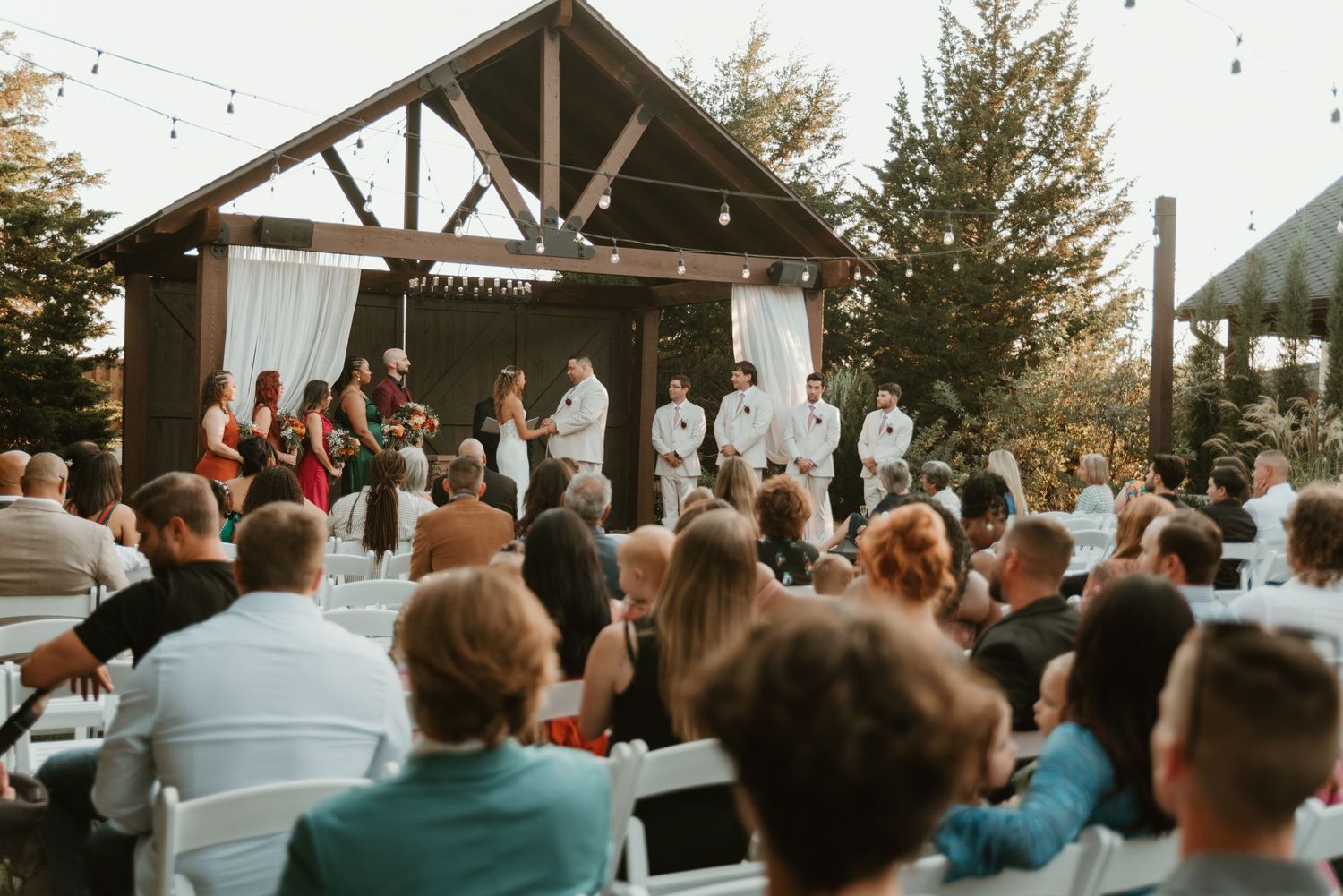 Wedding guests seated outdoors watching ceremony under a wooden pavilion surrounded by tall trees.