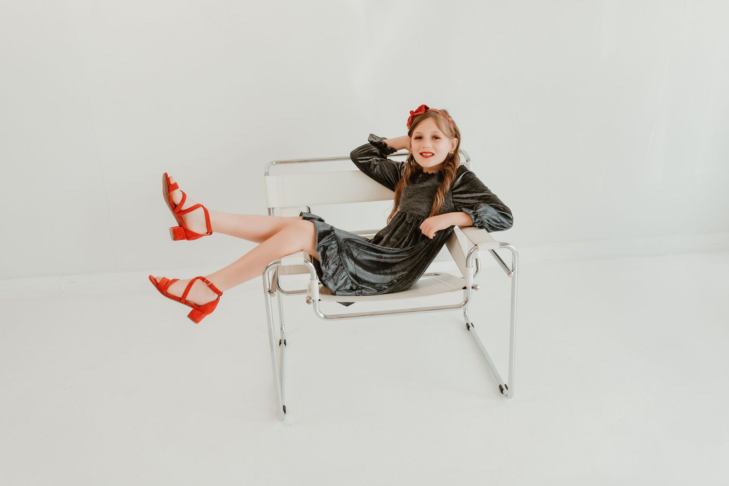Stylish young girl in velvet dress and red heels lounging in modern chrome chair on white backdrop.