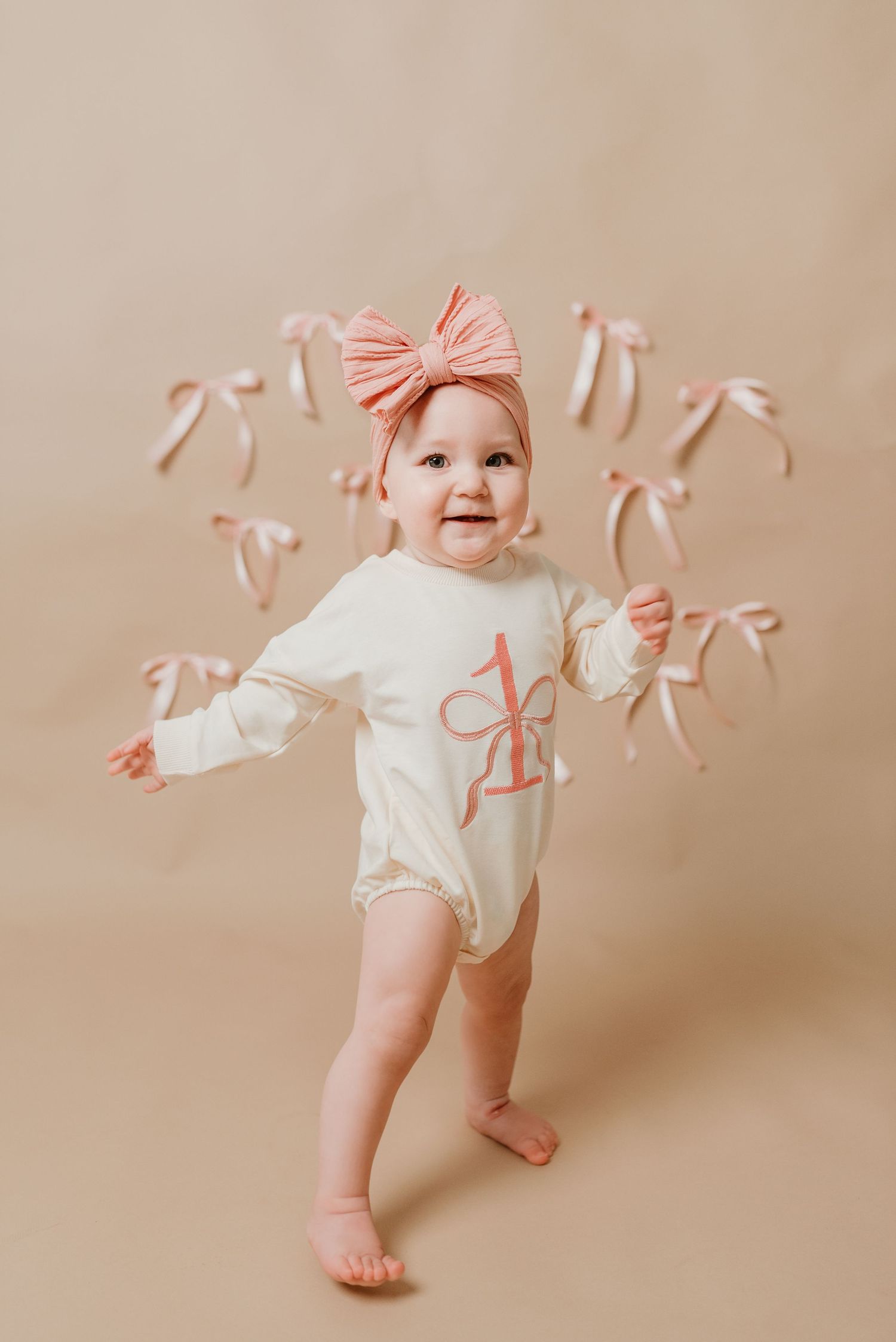 Happy one year old baby girl standing in cream bodysuit with pink initial surrounded by pink ribbon bows.
