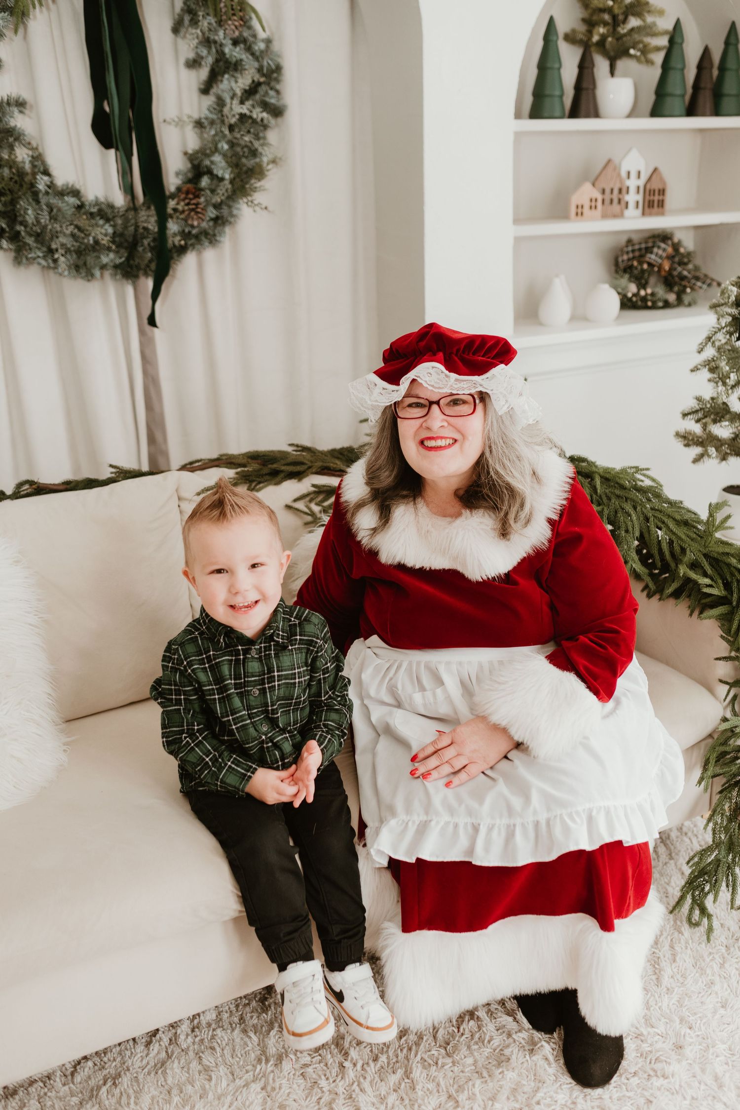 A young boy in green plaid sits smiling beside Mrs. Claus in a festive red outfit on a white couch.