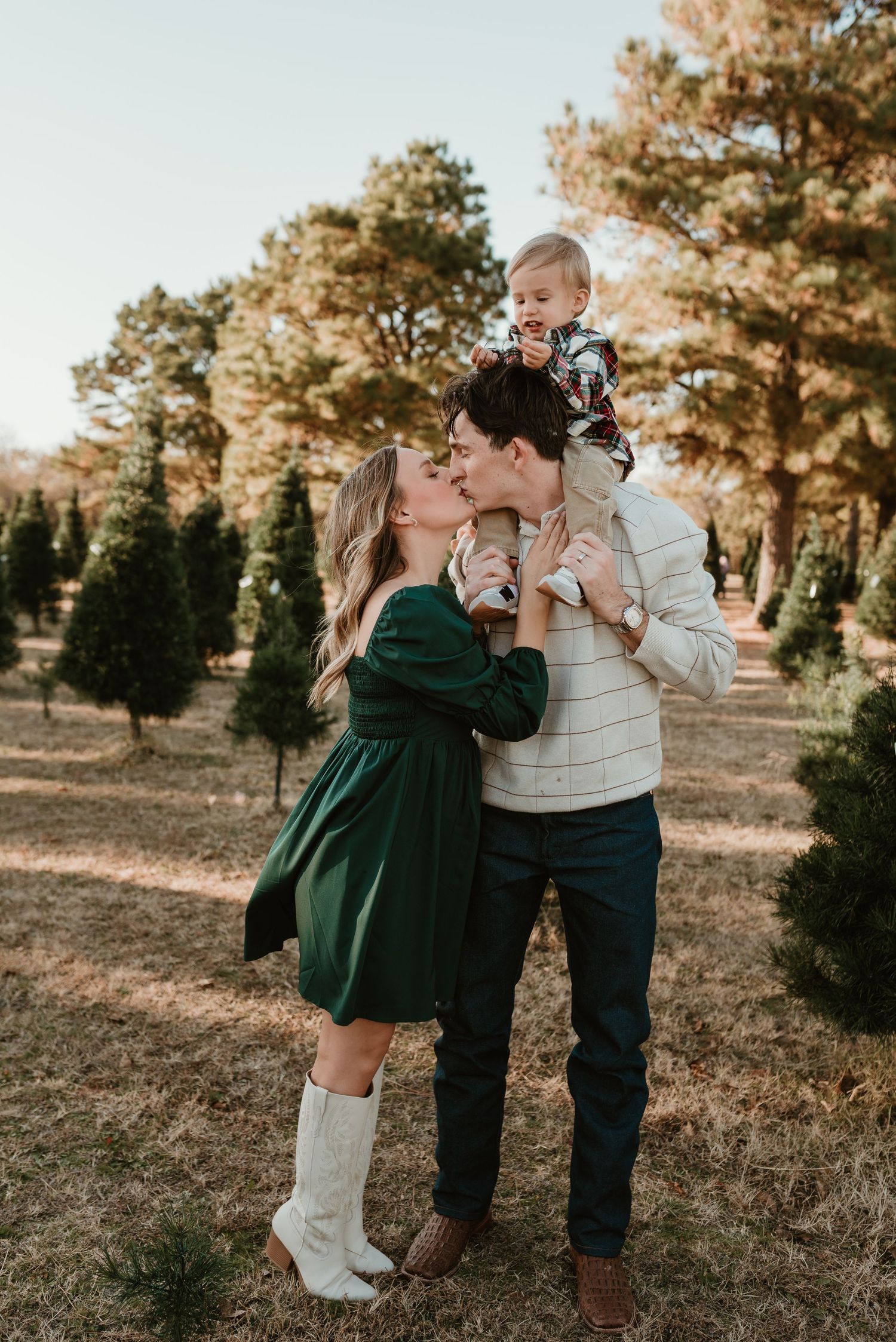 Young couple kisses while dad holds baby aloft at a scenic Christmas tree farm.