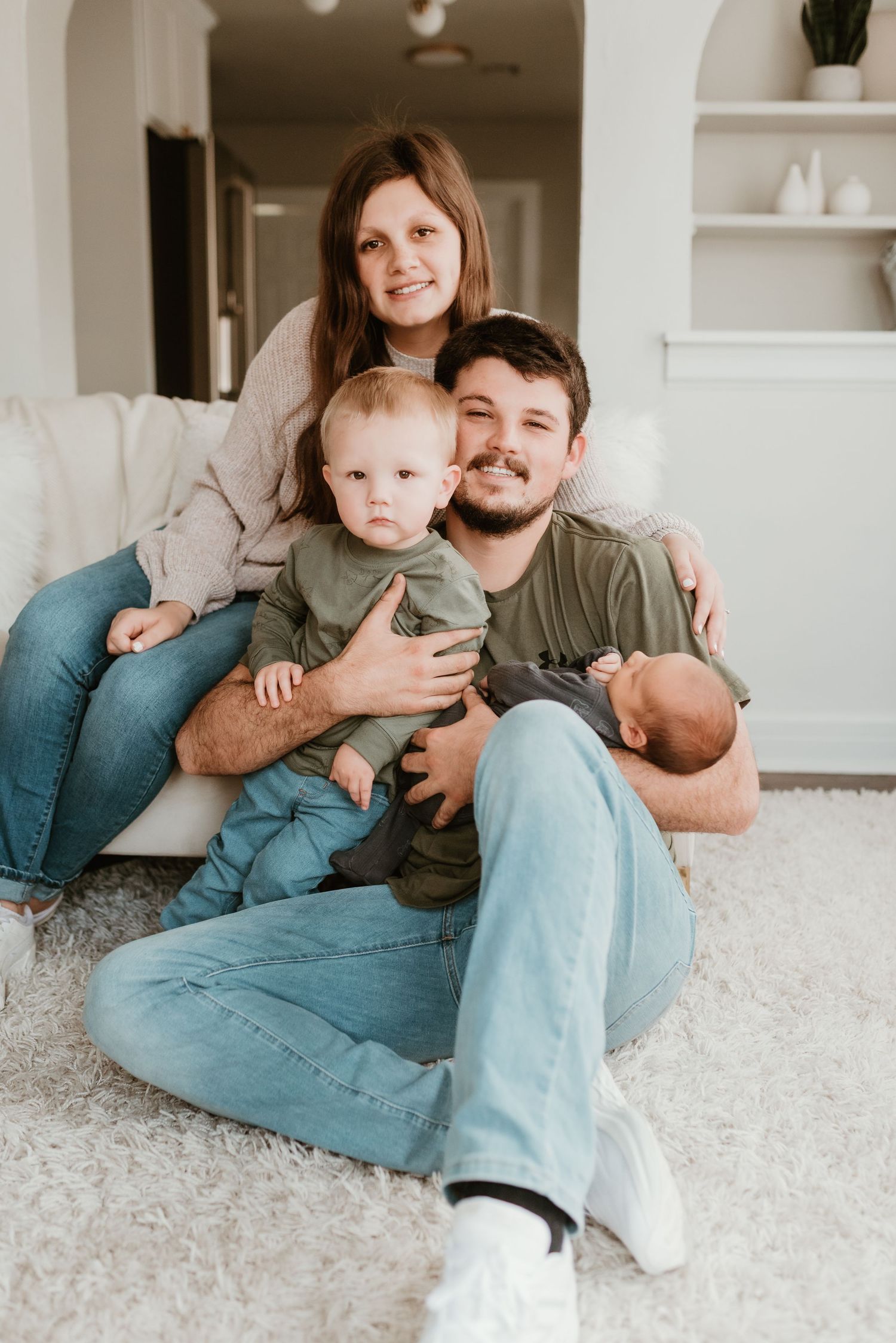 Young family with two babies poses together on living room floor, smiling warmly at camera.