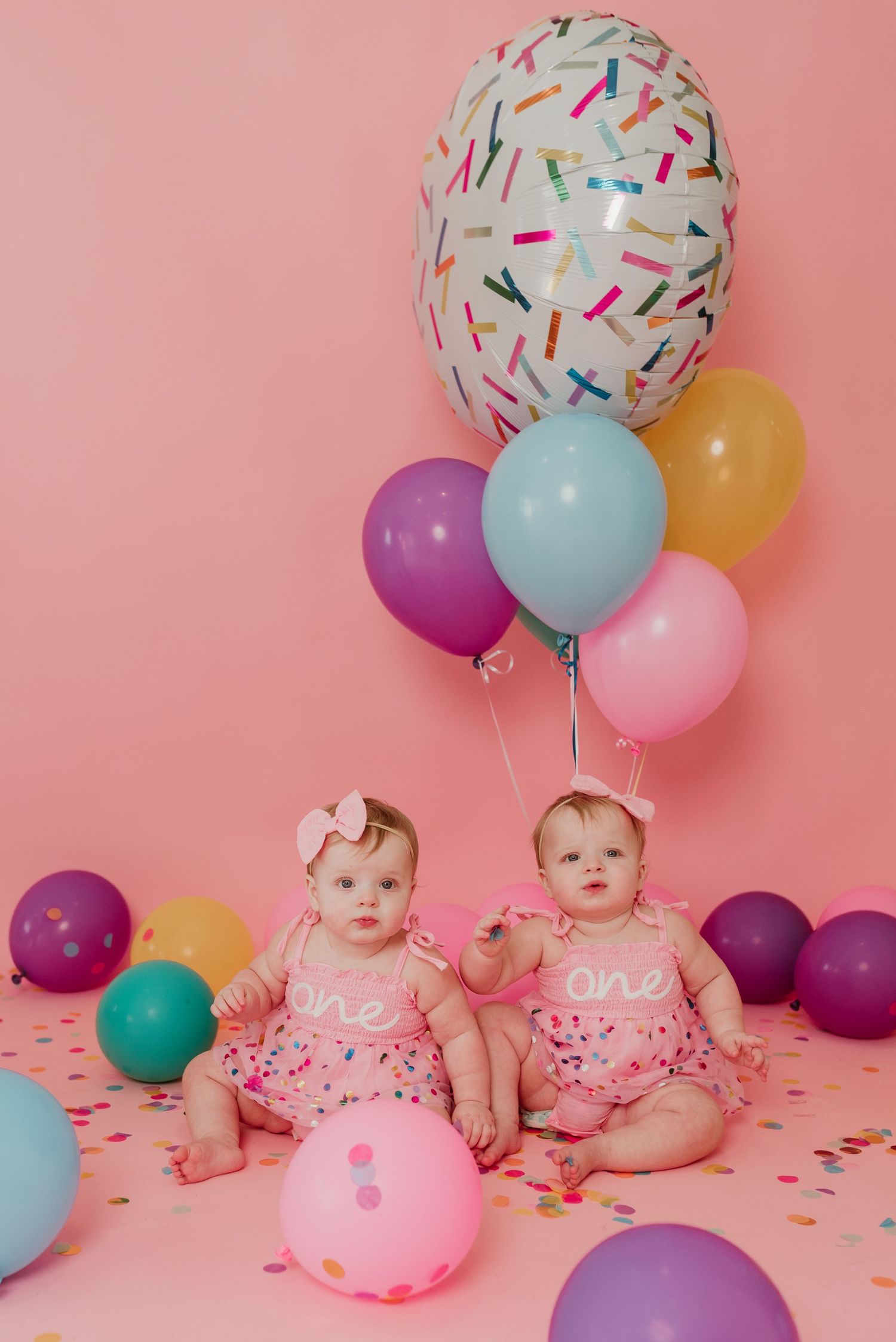 Twin baby girls in matching pink one birthday outfits sitting together surrounded by colorful balloons.