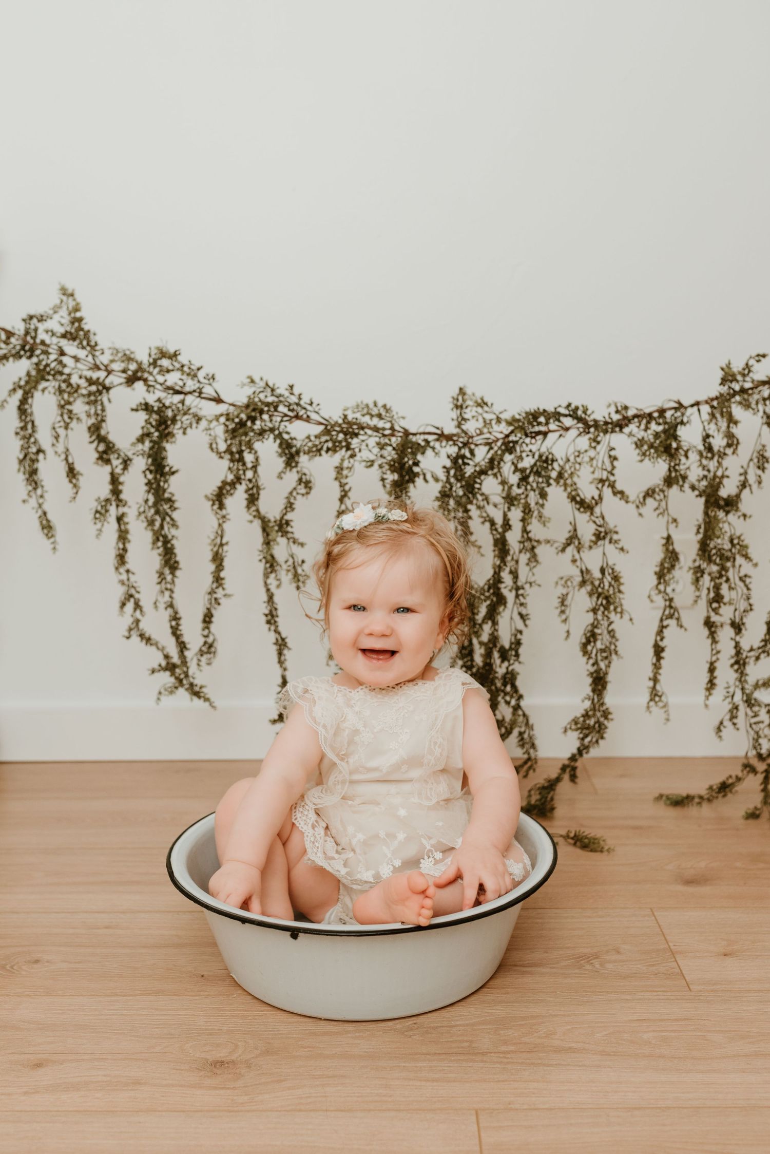Happy baby girl in lace romper sitting in enamel tub with floral garland backdrop for milestone photo.