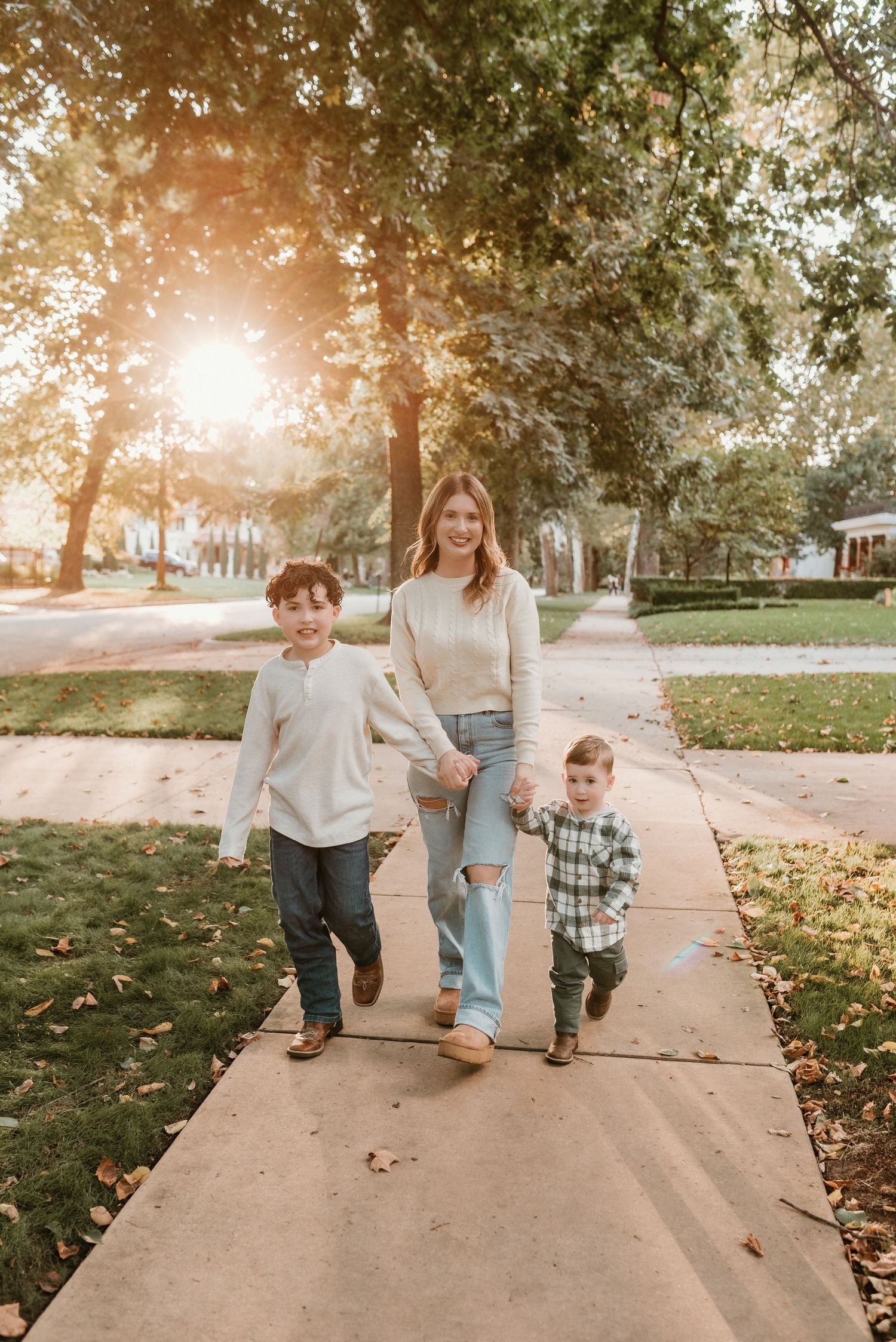 Young mother walks with two sons on sun-drenched sidewalk during golden hour family photos.