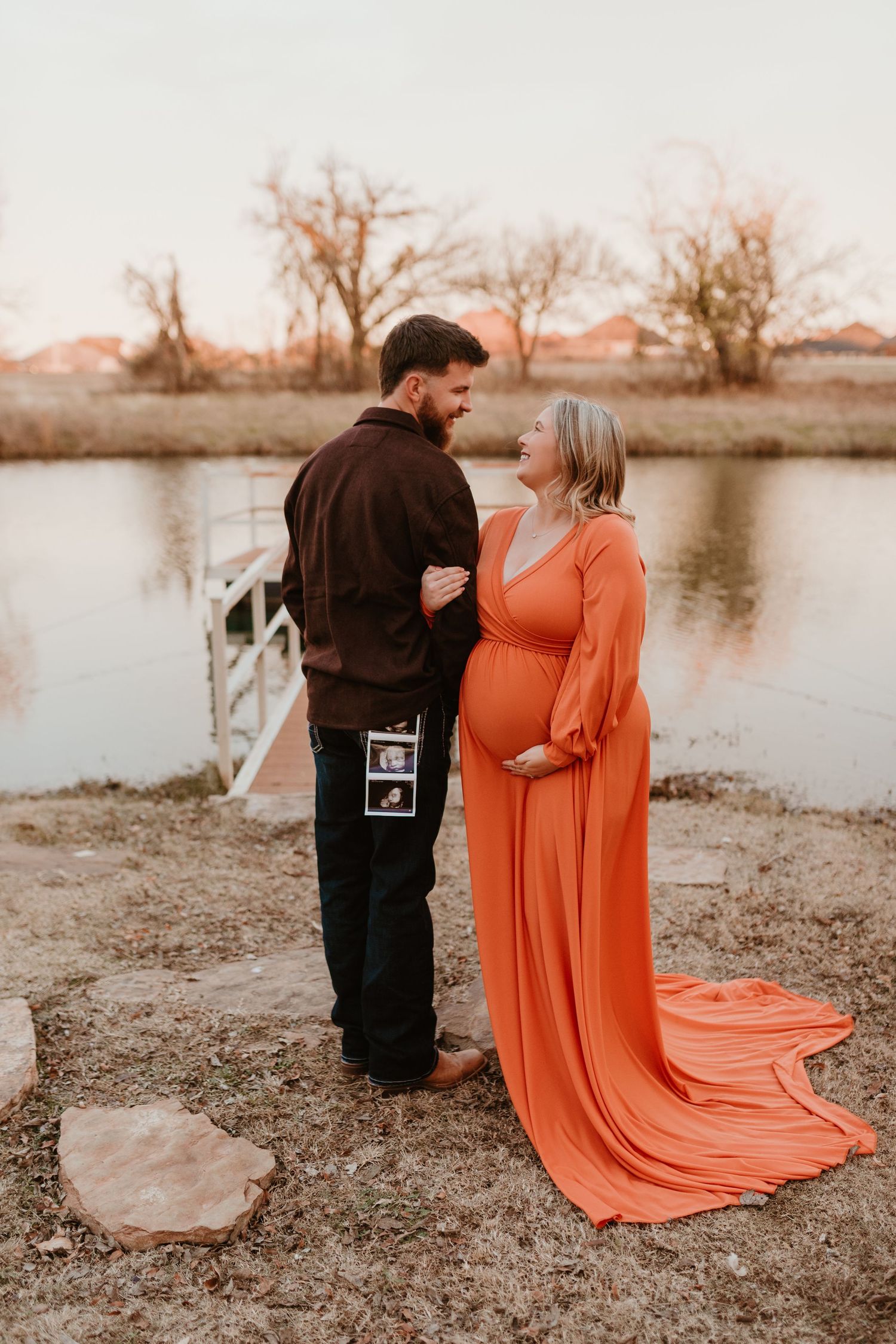 Expecting couple stands by pond holding ultrasound photos, woman in elegant orange maternity dress.