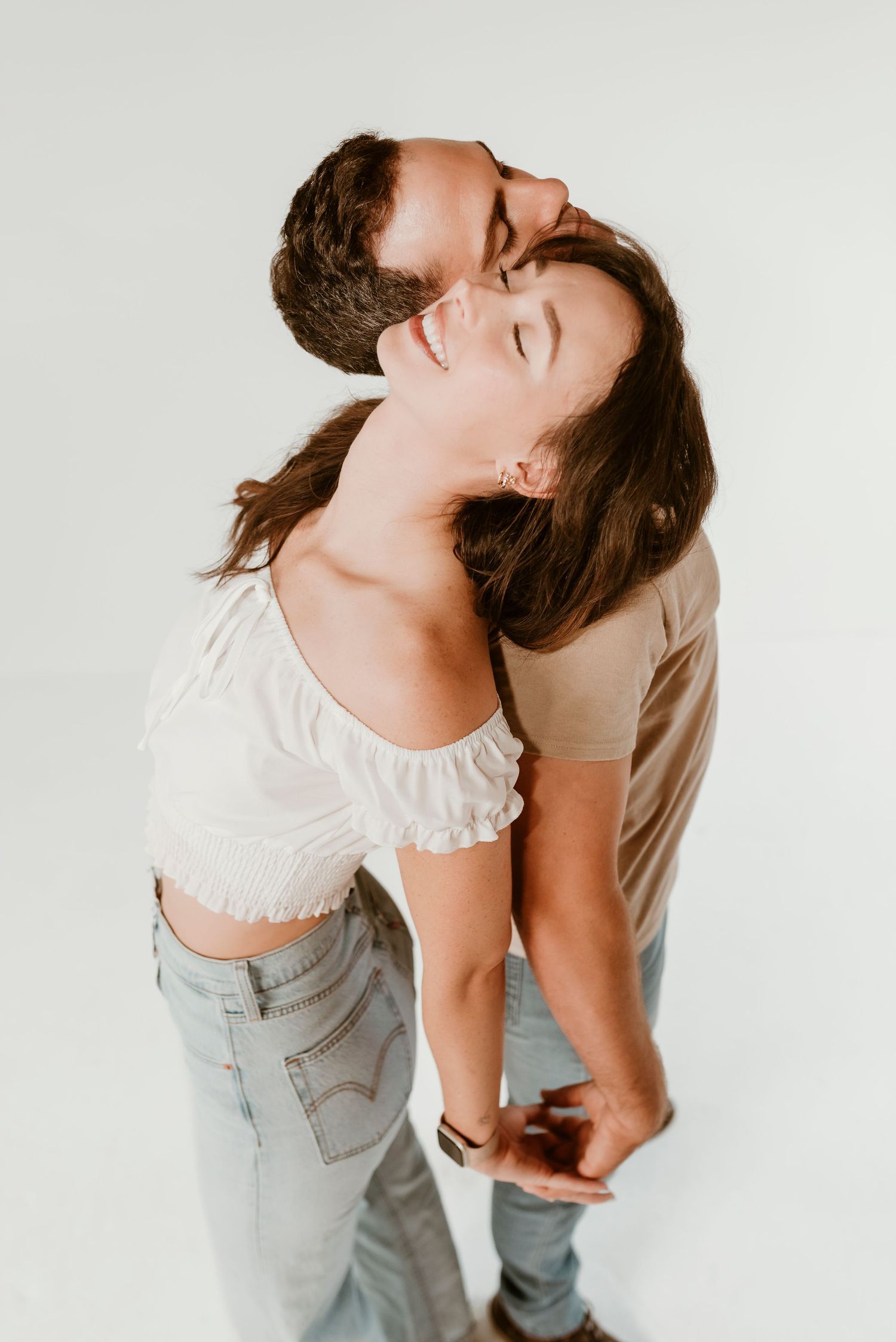 Man embracing woman from behind as she laughs joyfully in a white studio setting.