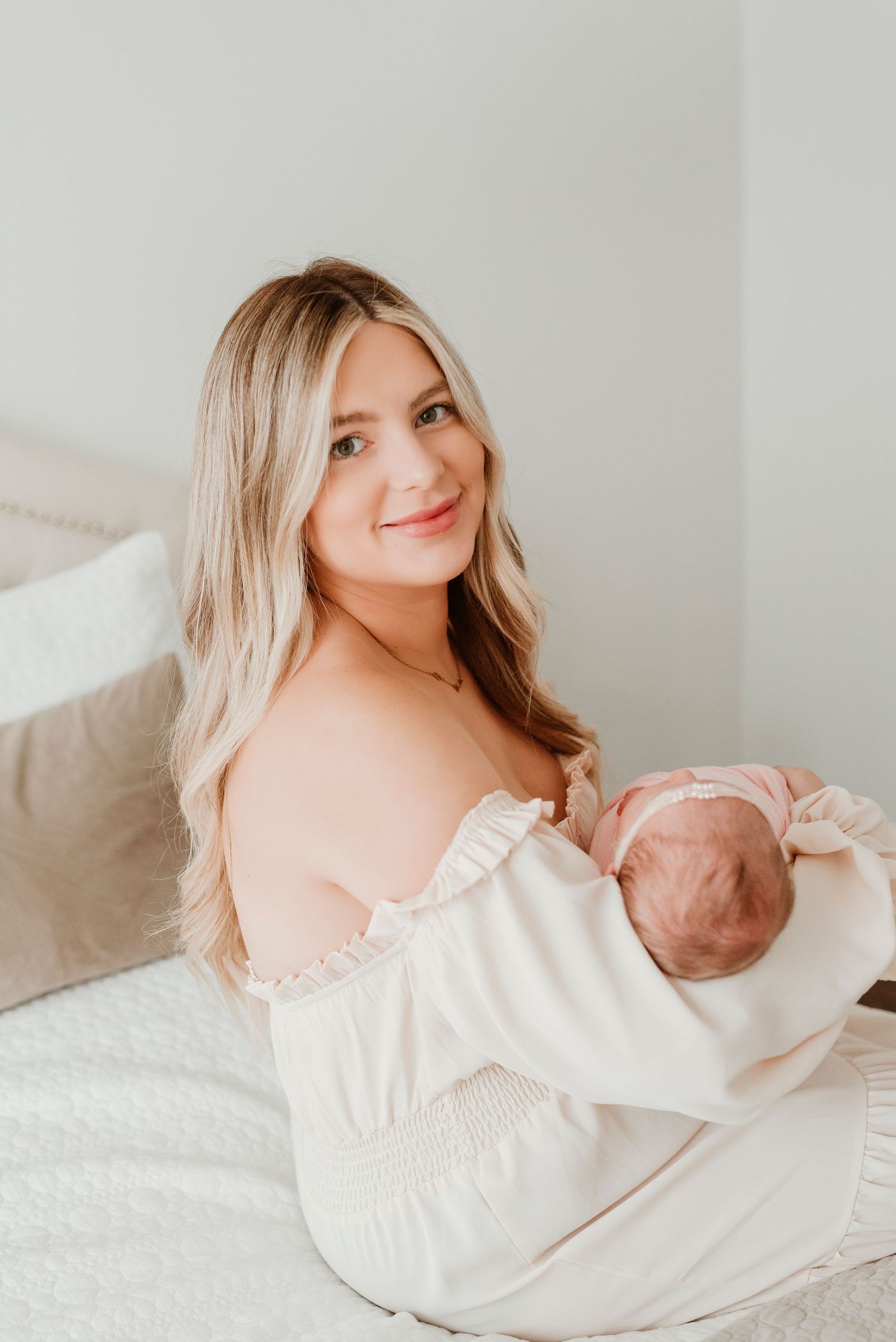 A smiling blonde mother in a white off-shoulder top sits on a bed, holding her newborn baby close.