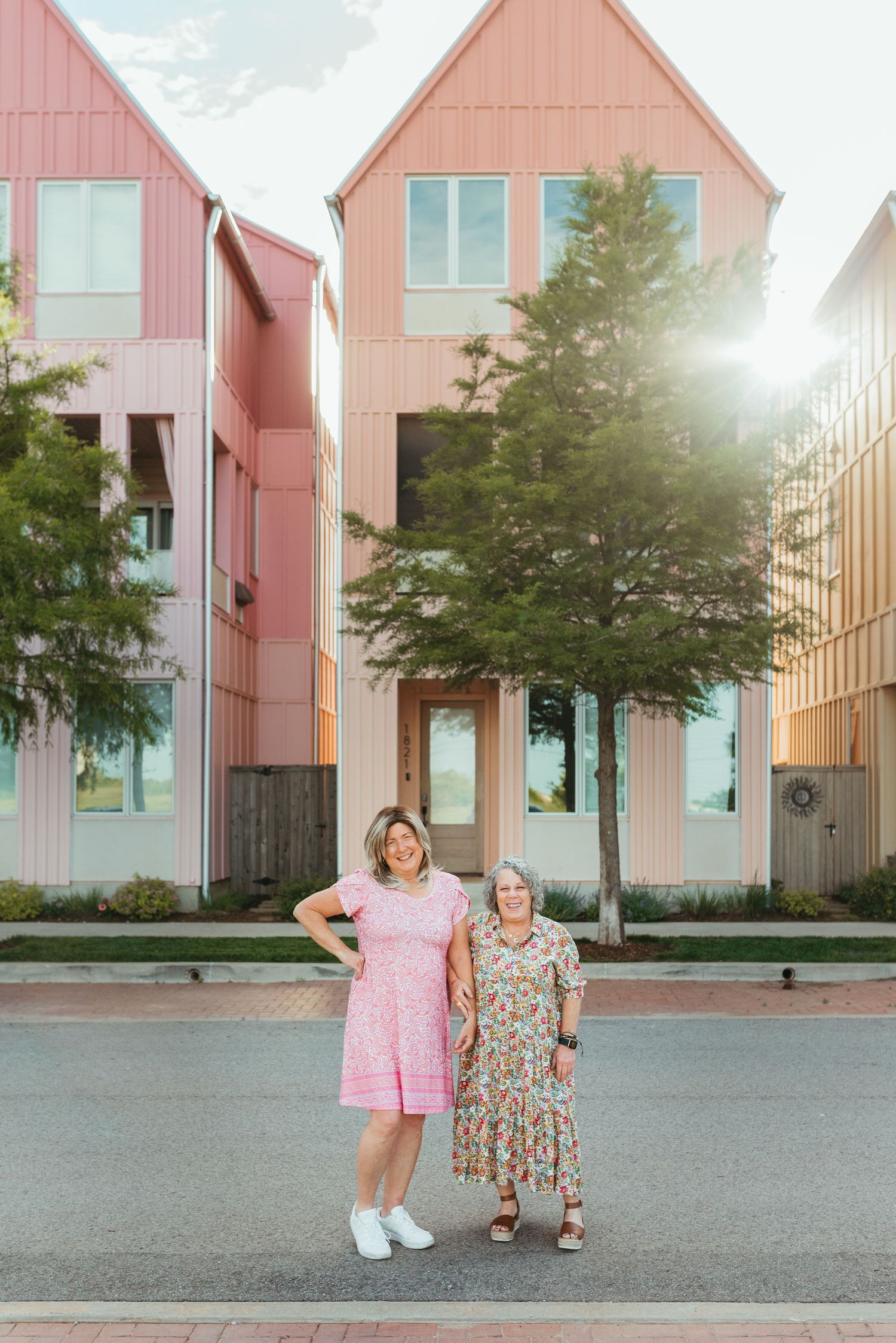 Two women in pink and floral dresses posing together on a sunny street with colorful modern townhomes.