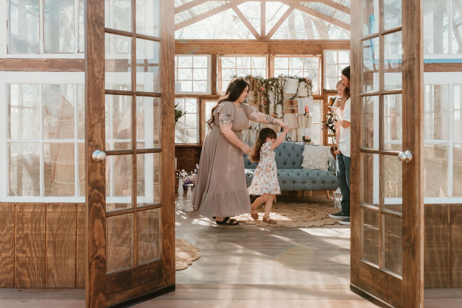 Family gathering in sunlit wood-framed greenhouse studio with French doors, mother dancing with toddler.