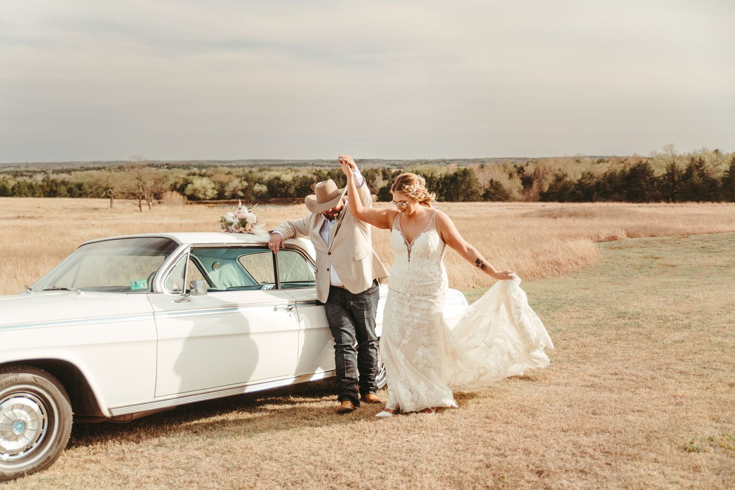 Bride and groom dance joyfully beside a vintage white classic car in a golden countryside field.