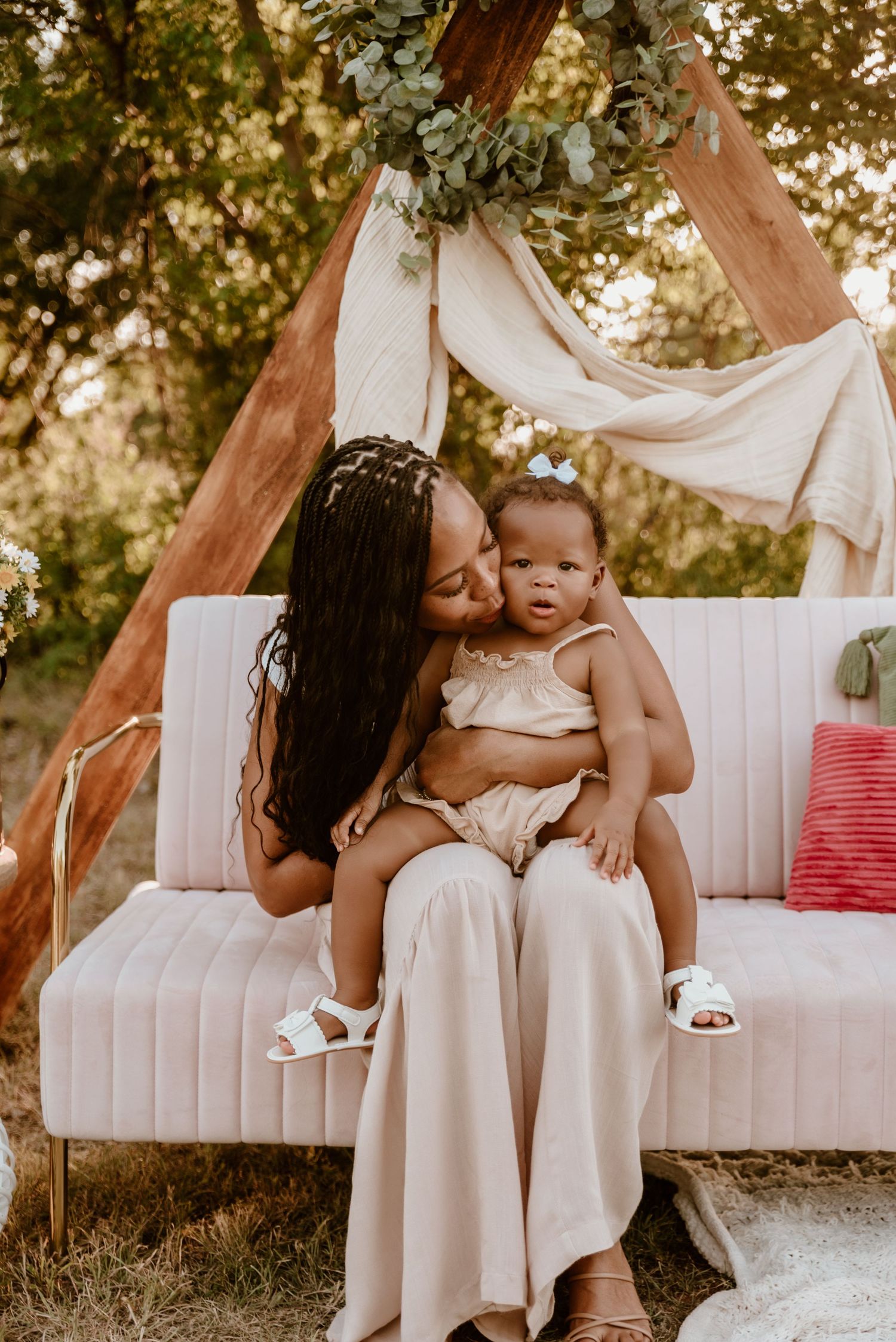 A mother tenderly kisses her baby girl while seated on a pink sofa in a boho outdoor setting.