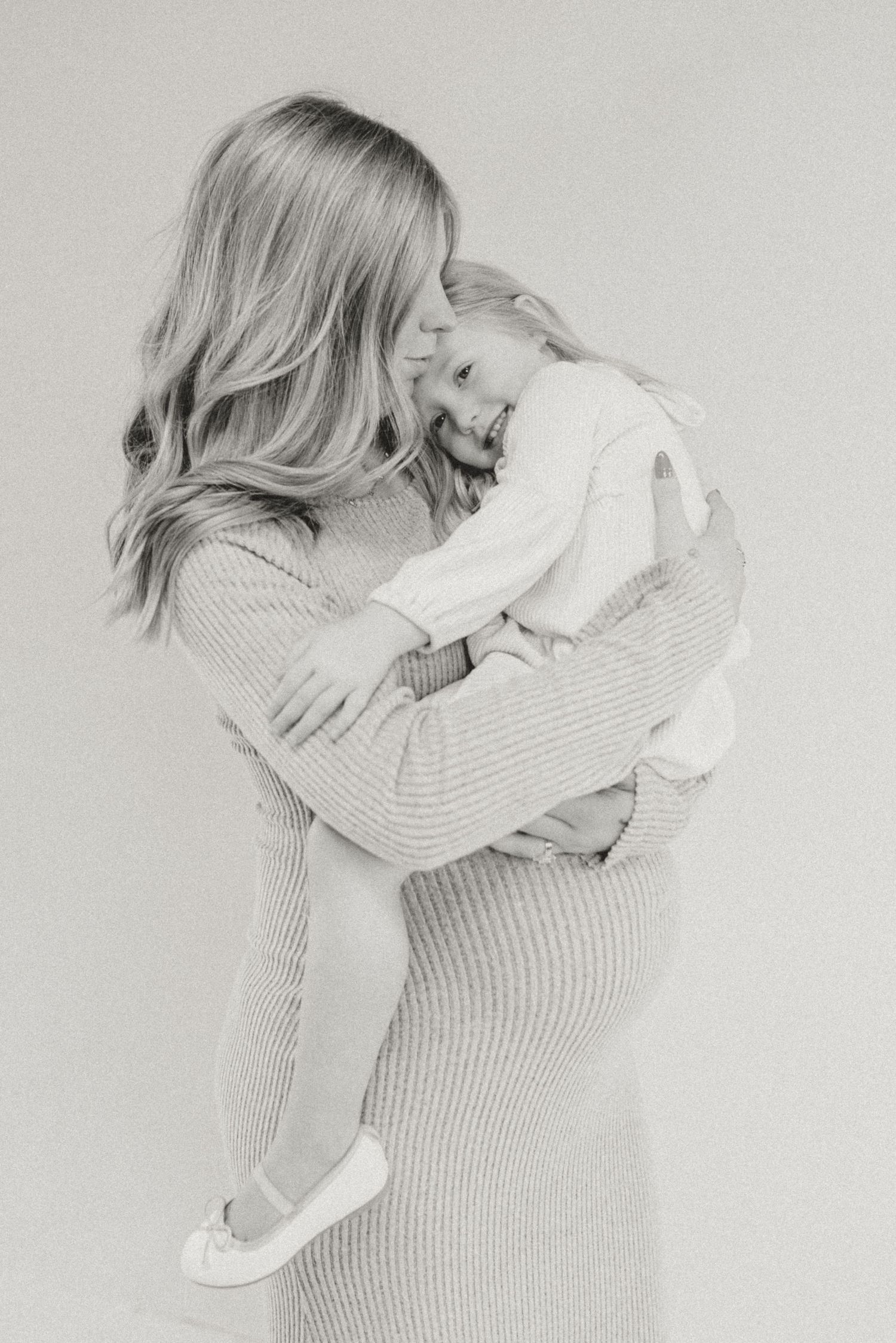 Black and white photo of pregnant mother in knit dress hugging smiling toddler tenderly.