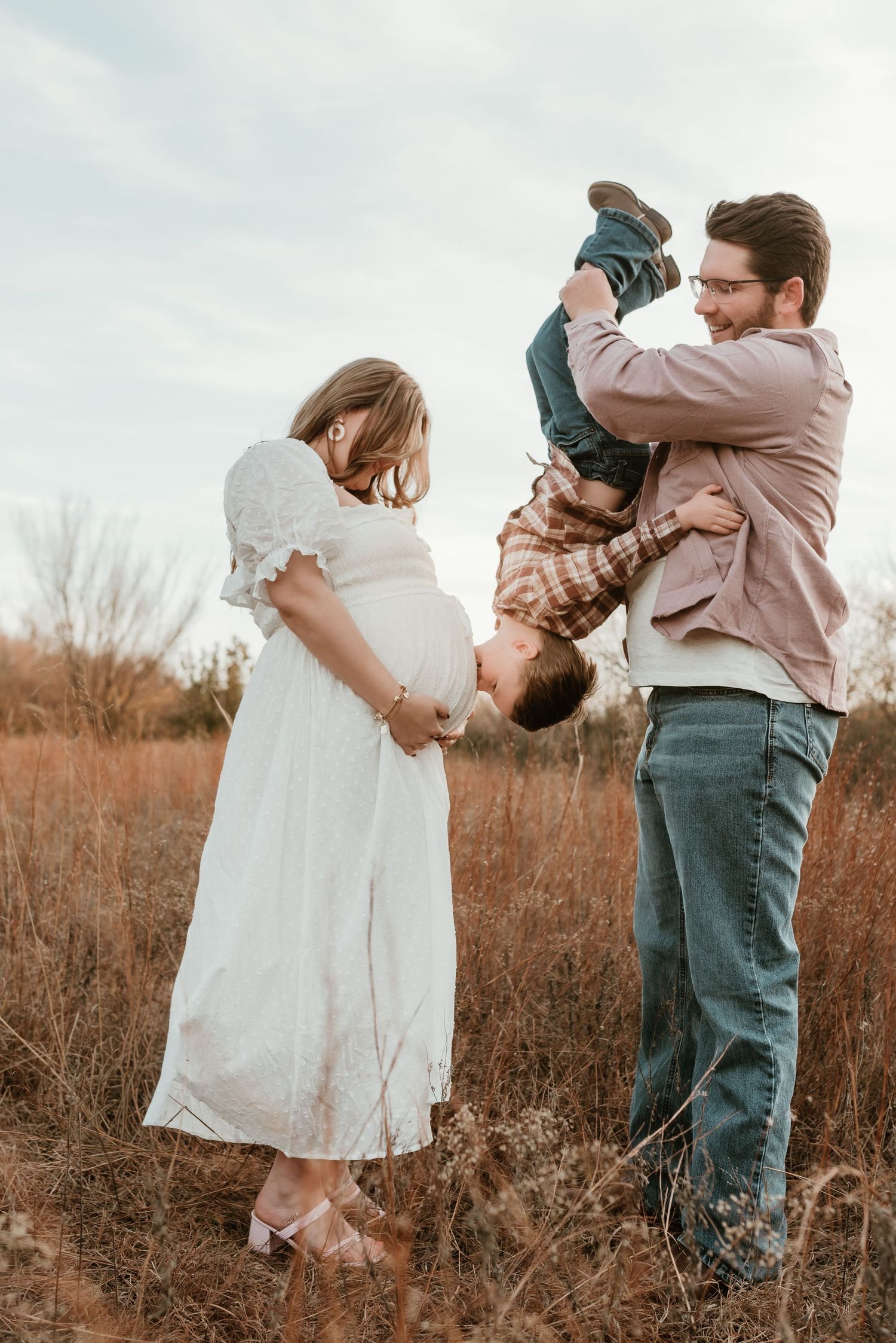 Smiling dad flips giggling toddler upside down while pregnant mom watches in autumn field.
