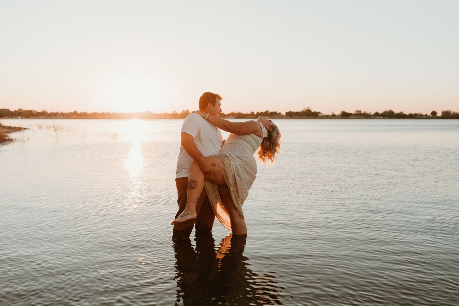 Man dipping woman in shallow lake water during a beautiful golden sunset.