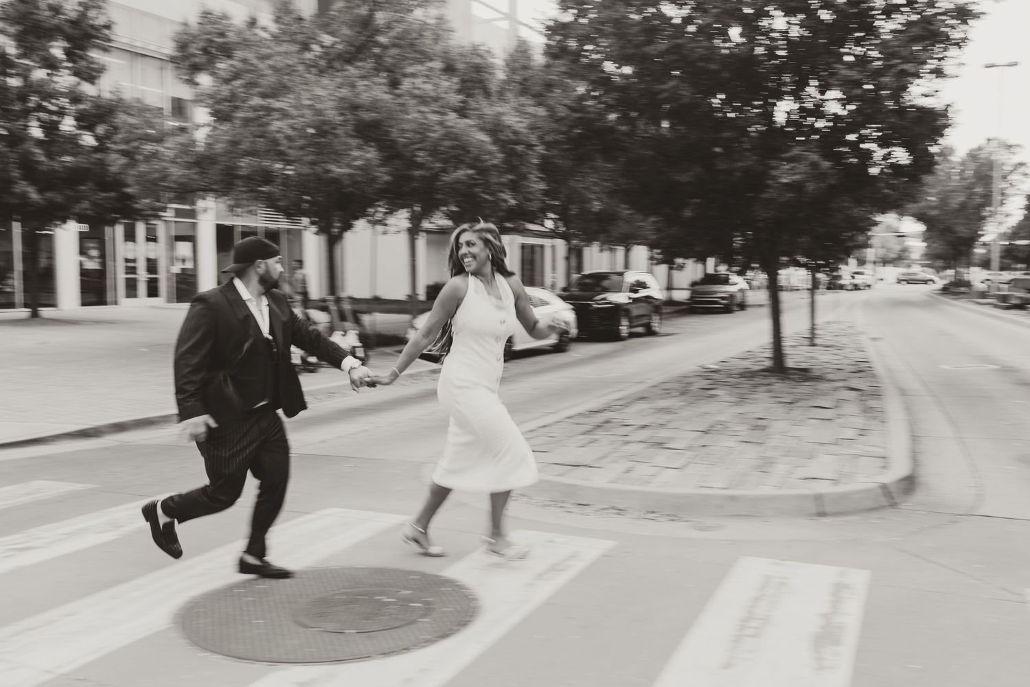Black and white photo of a couple in wedding attire running hand in hand across a city crosswalk.