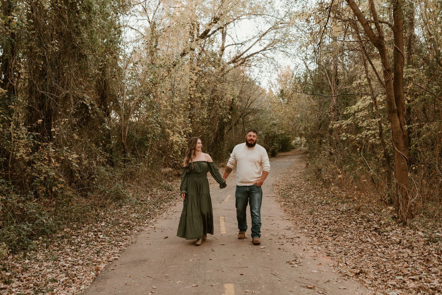 Couple holds hands walking down a scenic tree-lined road in fall foliage setting.