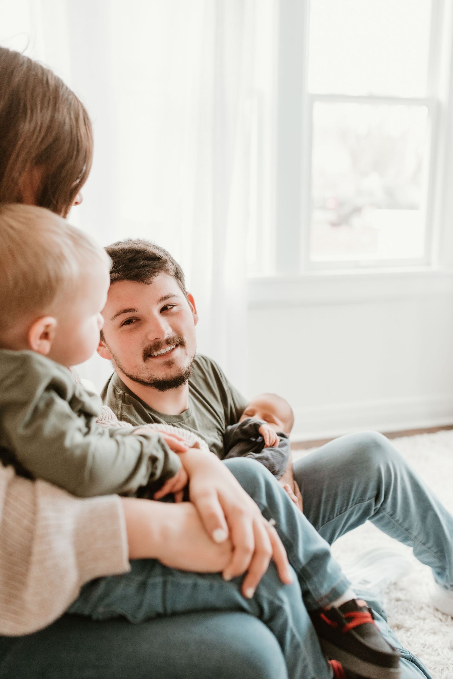 Smiling father sitting on floor holding toddler and newborn with mother beside bright window.