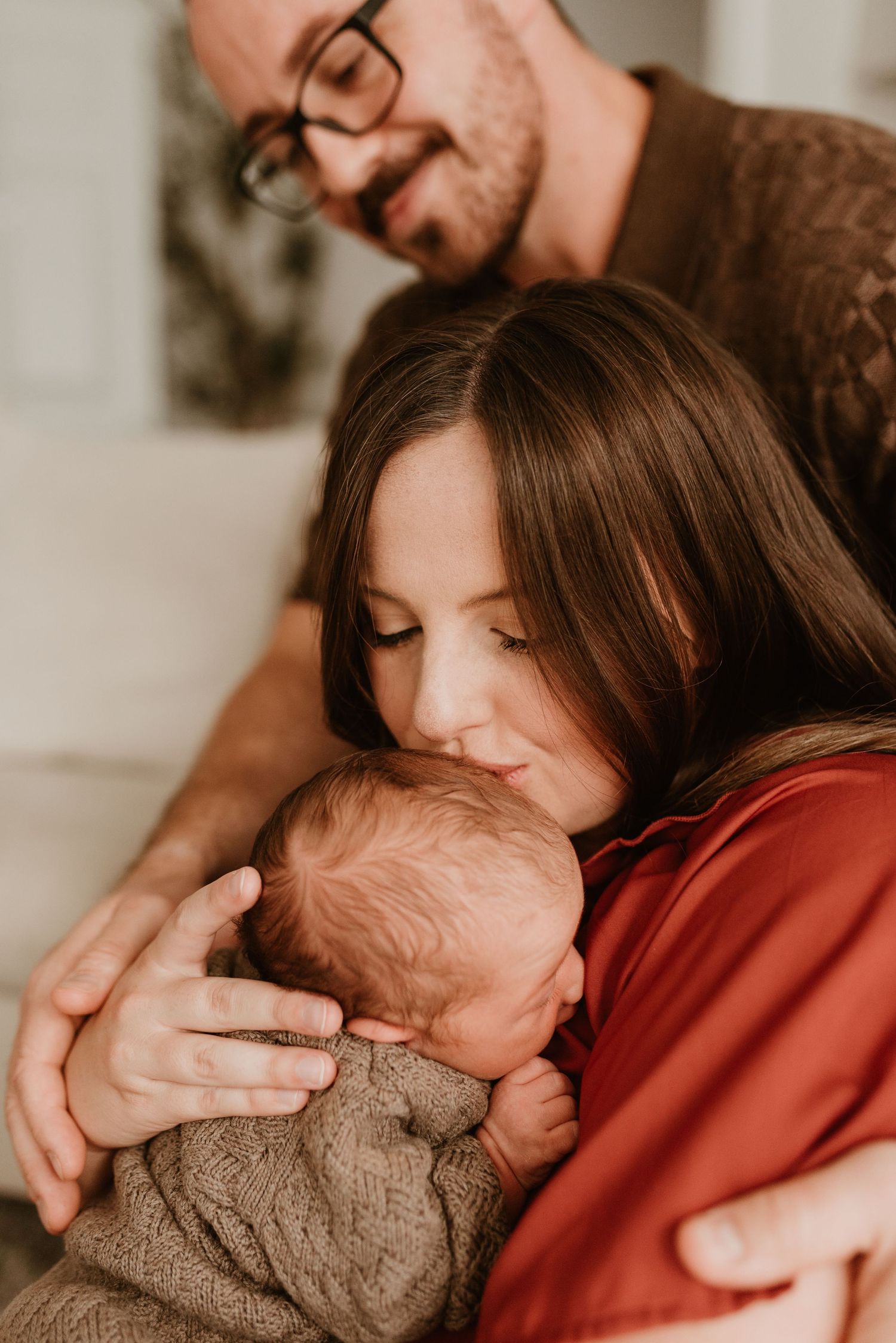 A mother in a red dress tenderly kisses her newborn while the father in glasses looks on lovingly from behind.