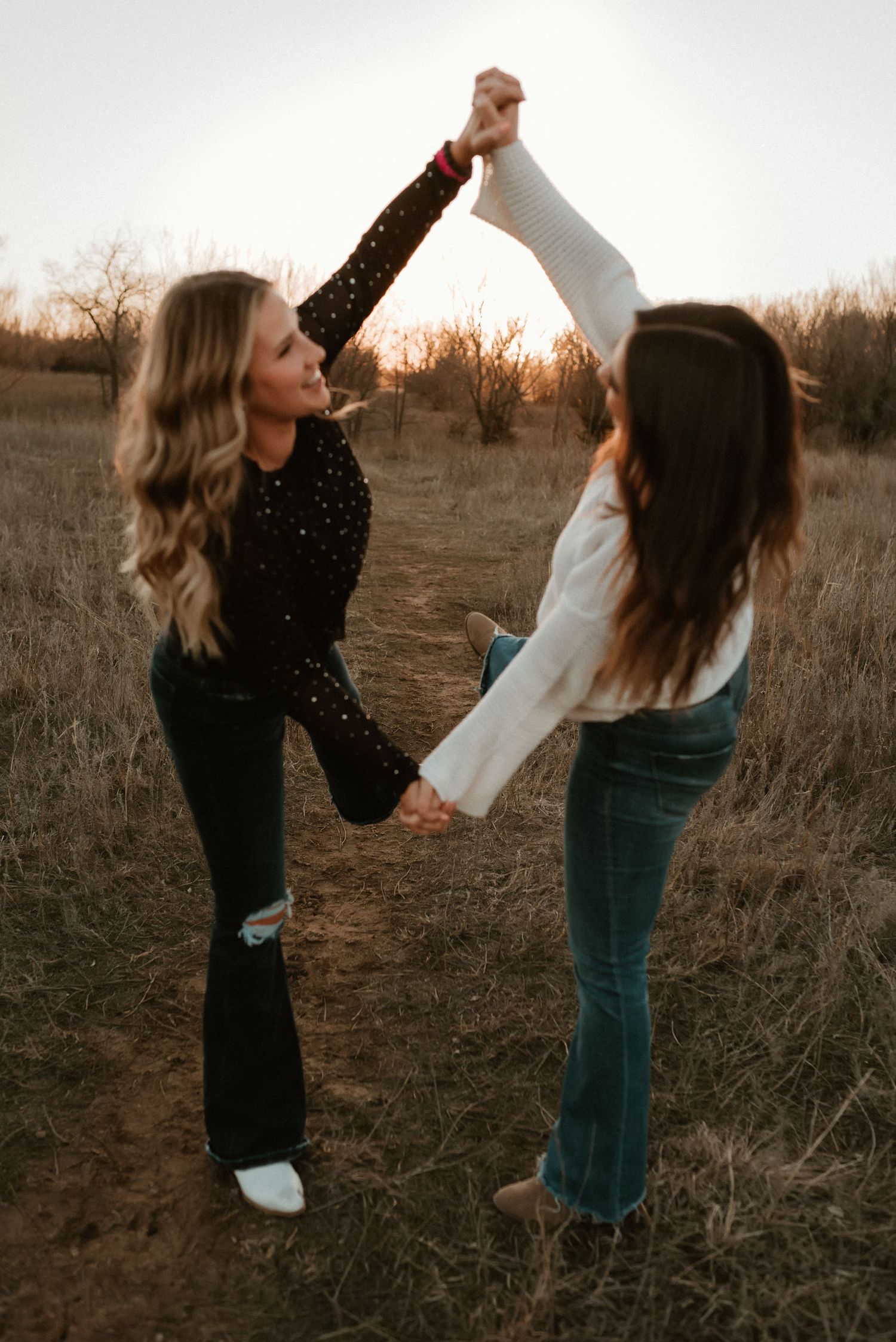 Two young women dance together holding hands in a golden field at sunset wearing boho-style outfits.