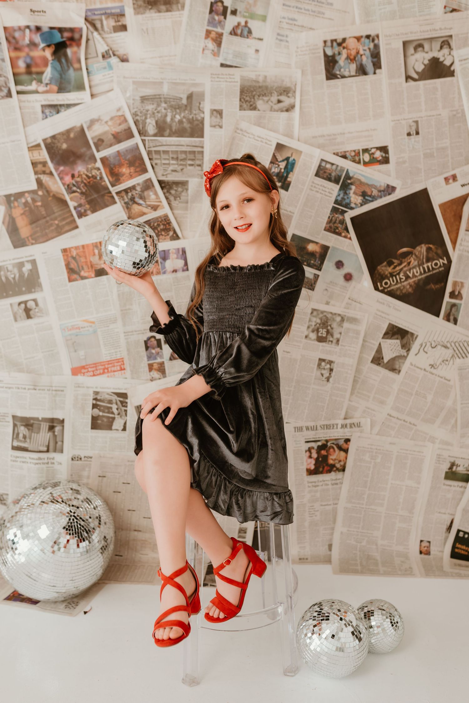 Young girl in velvet dress holding disco ball posing before newspaper backdrop with mirror ball decor.