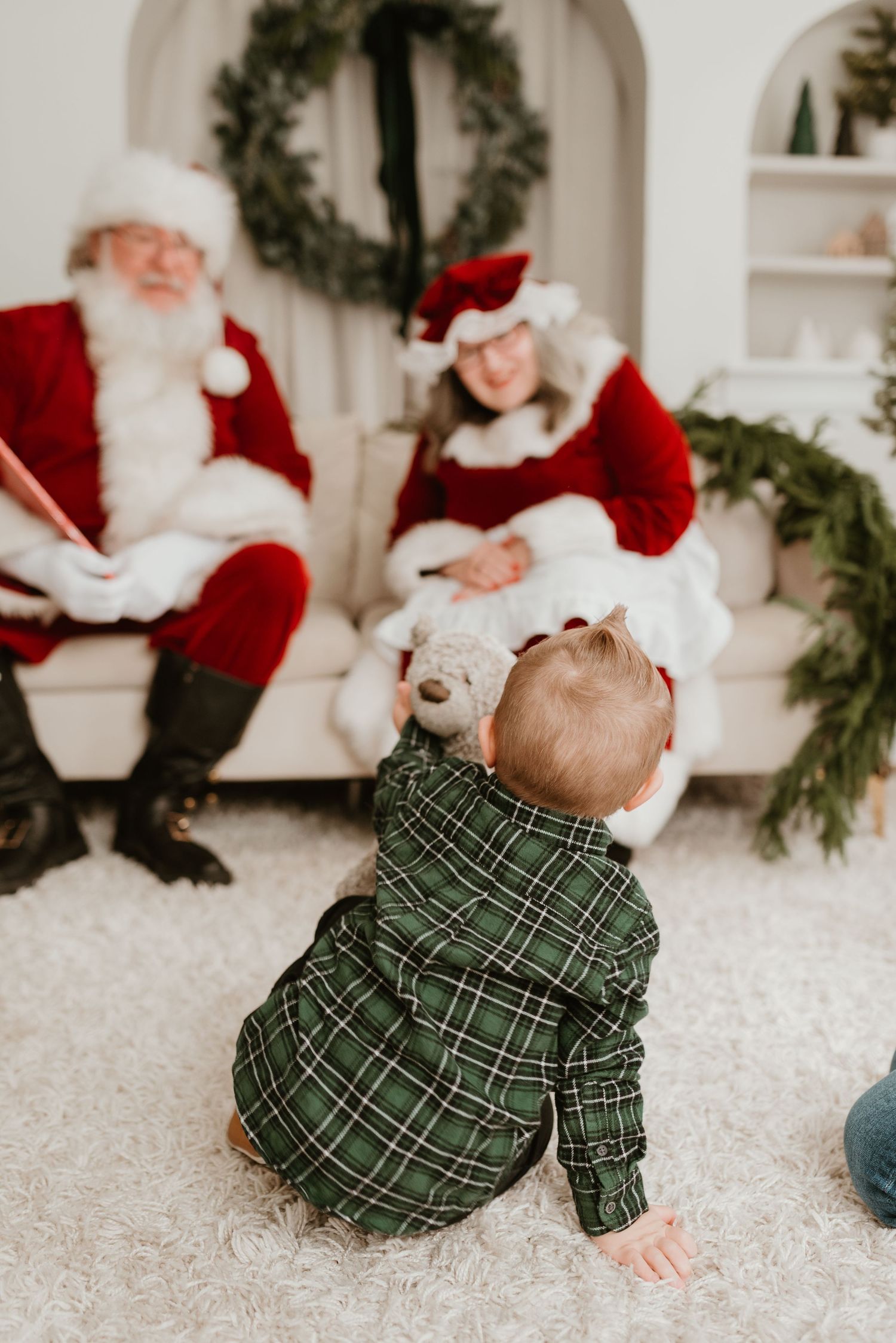 A baby in green plaid crawls toward Santa and Mrs. Claus seated on a white couch with holiday decor.