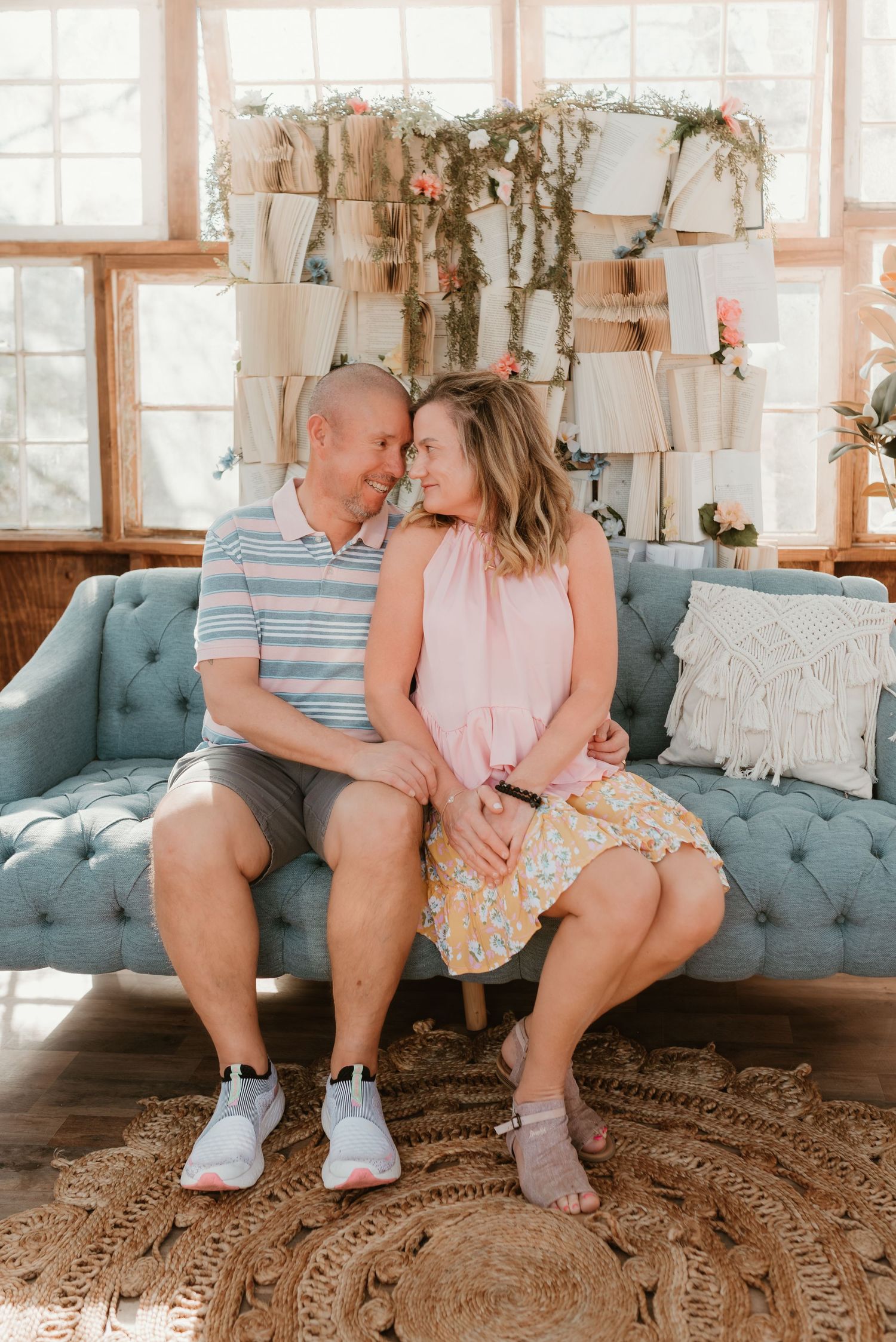 A couple sits closely on a tufted blue sofa with floral backdrop in a cozy indoor setting.