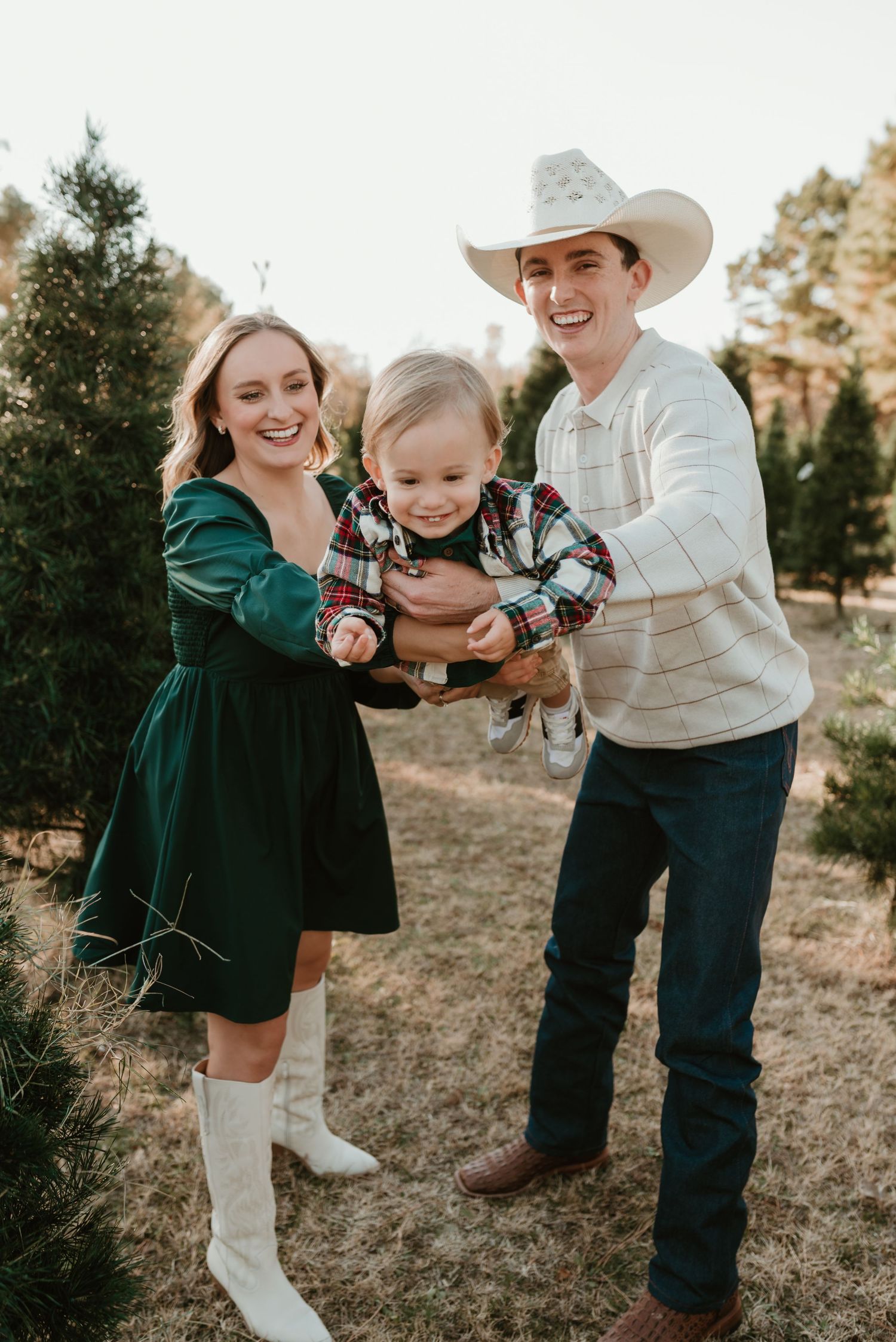 Laughing young family with baby poses together at a Christmas tree farm outdoors.