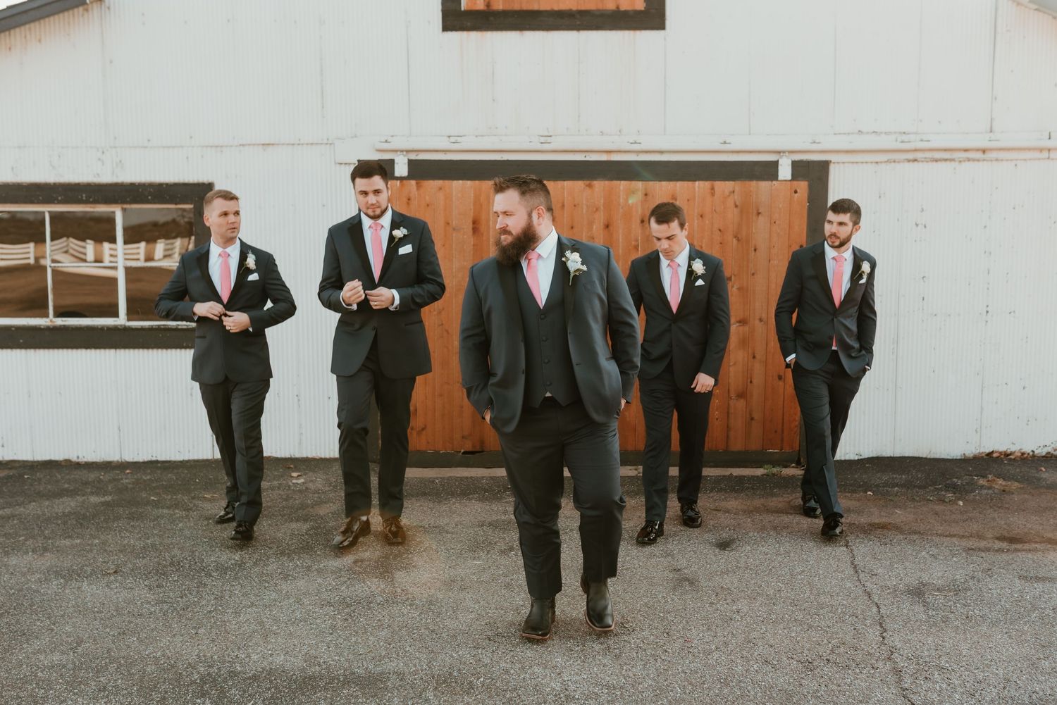 Five groomsmen in navy suits with pink ties walking confidently in front of a white barn.