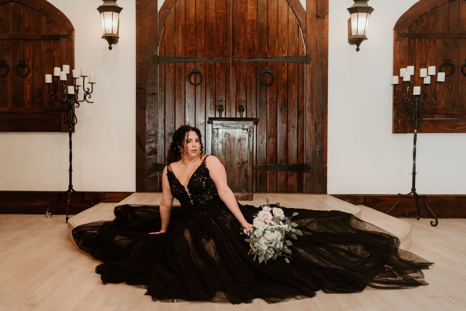 Bride in dramatic black lace wedding gown poses on floor with white bouquet before rustic wooden doors.