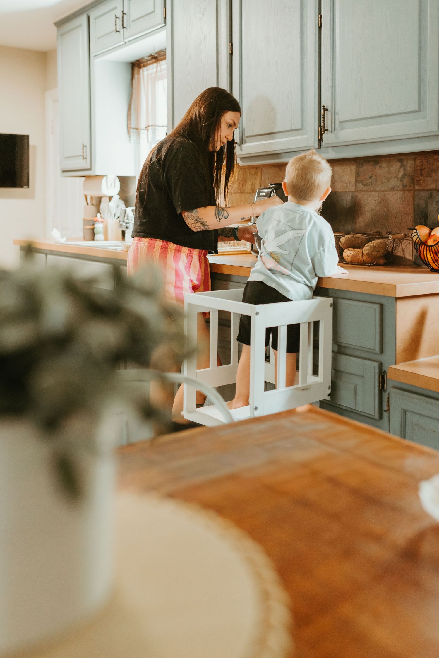 Mother helps toddler standing on a learning tower at a kitchen counter with gray cabinets.