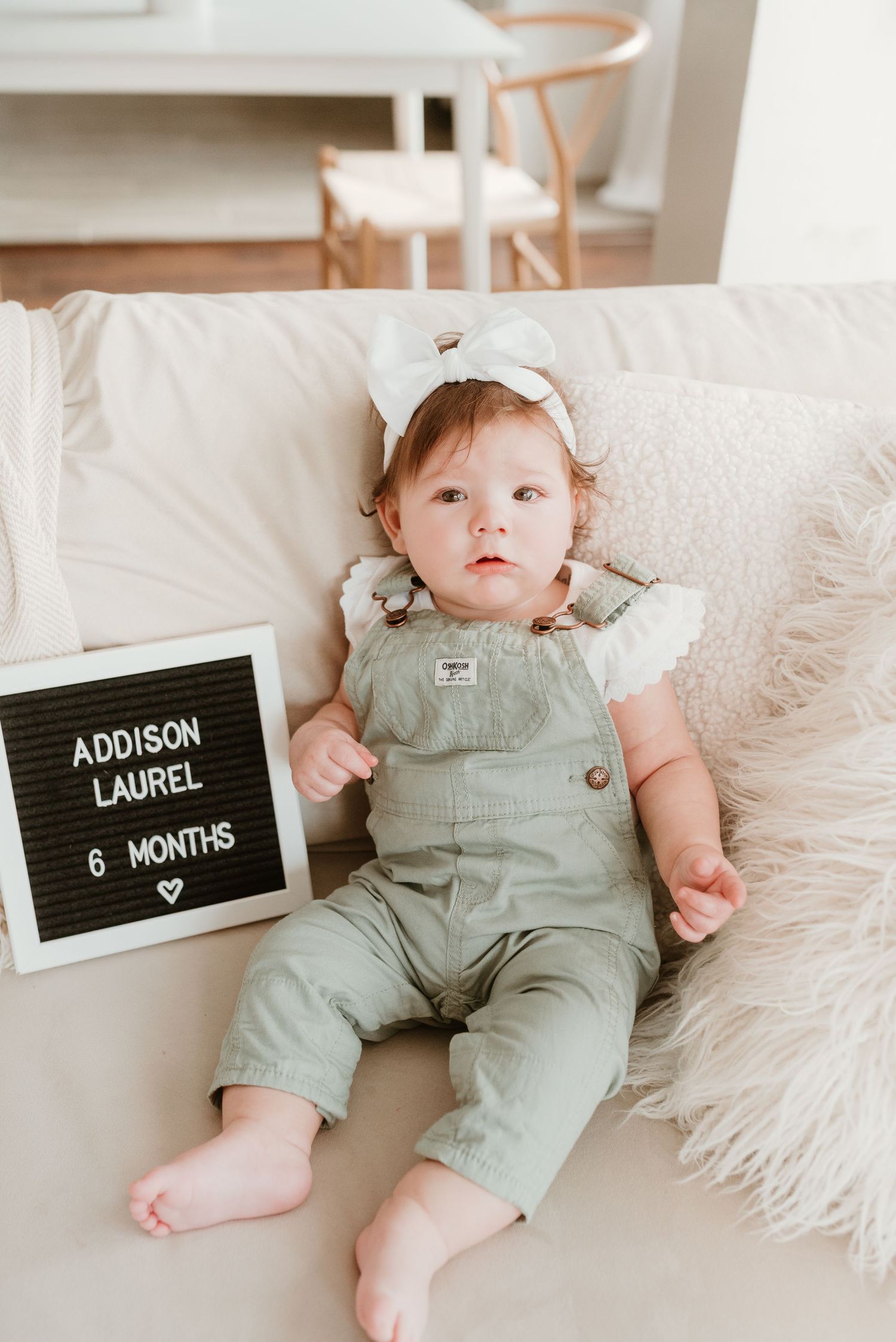 Baby girl Addison Laurel at 6 months old wearing green overalls and white bow headband posed on cream sofa.