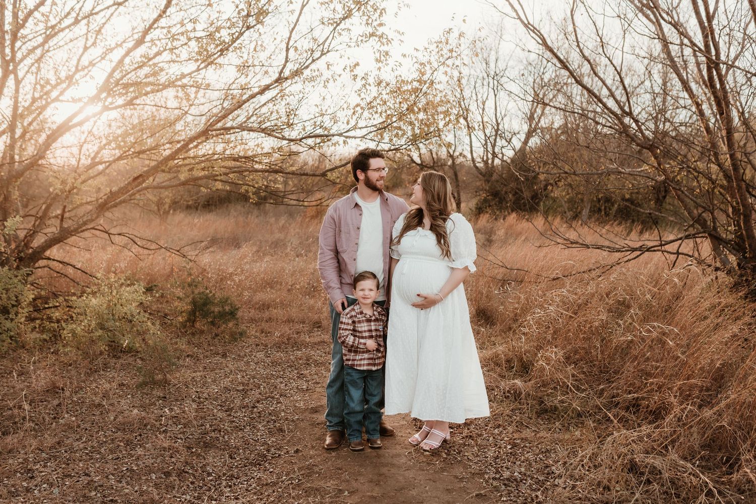 Expecting family with young boy standing in golden field at sunset, mother in white maternity dress.