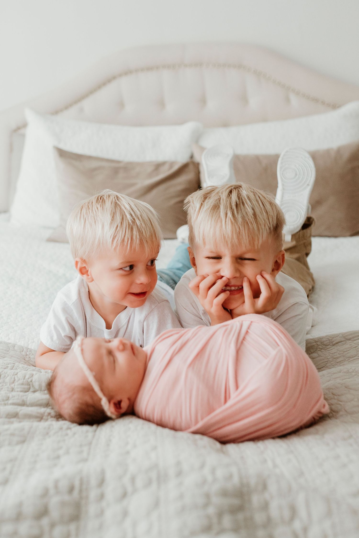 Two blonde toddler brothers smiling at swaddled newborn baby sister lying on a bed.