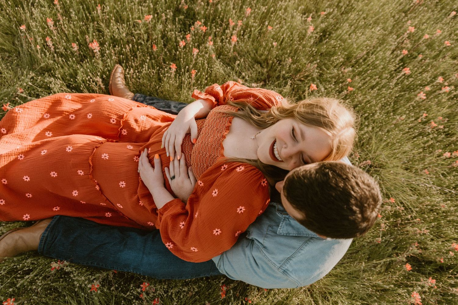 Couple lying in a wildflower meadow, woman in red floral dress smiling while embracing her partner.