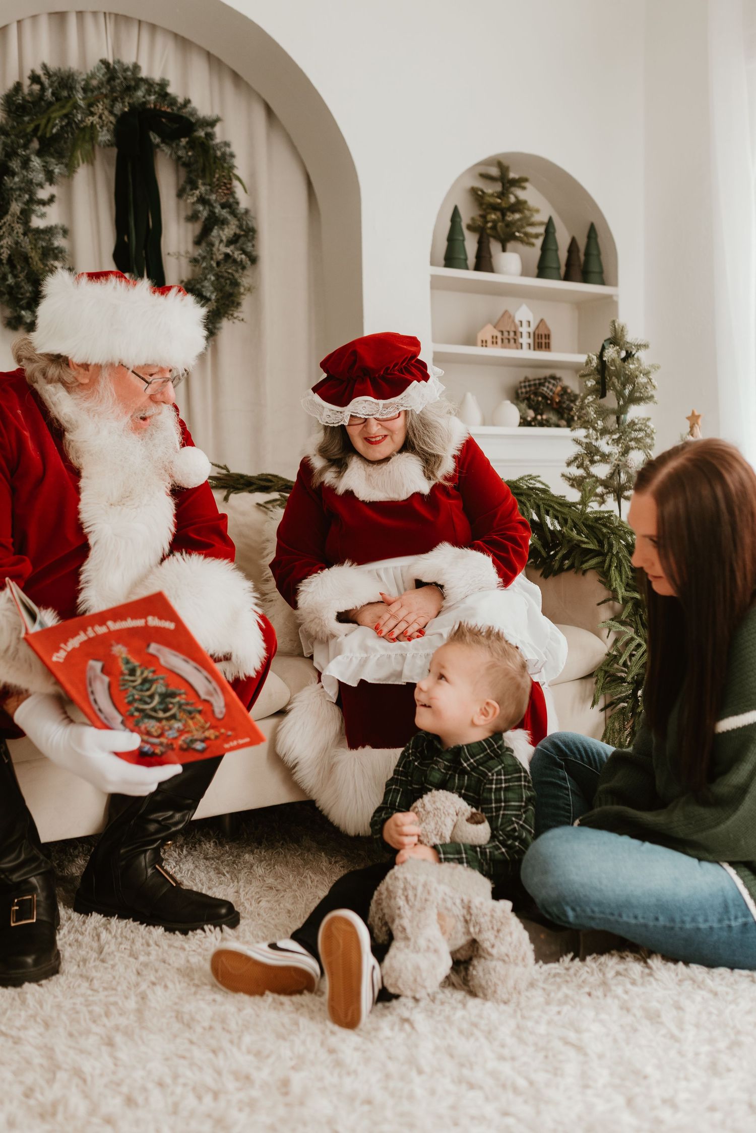 Santa reads a Christmas book to a toddler and mother while Mrs. Claus looks on warmly.