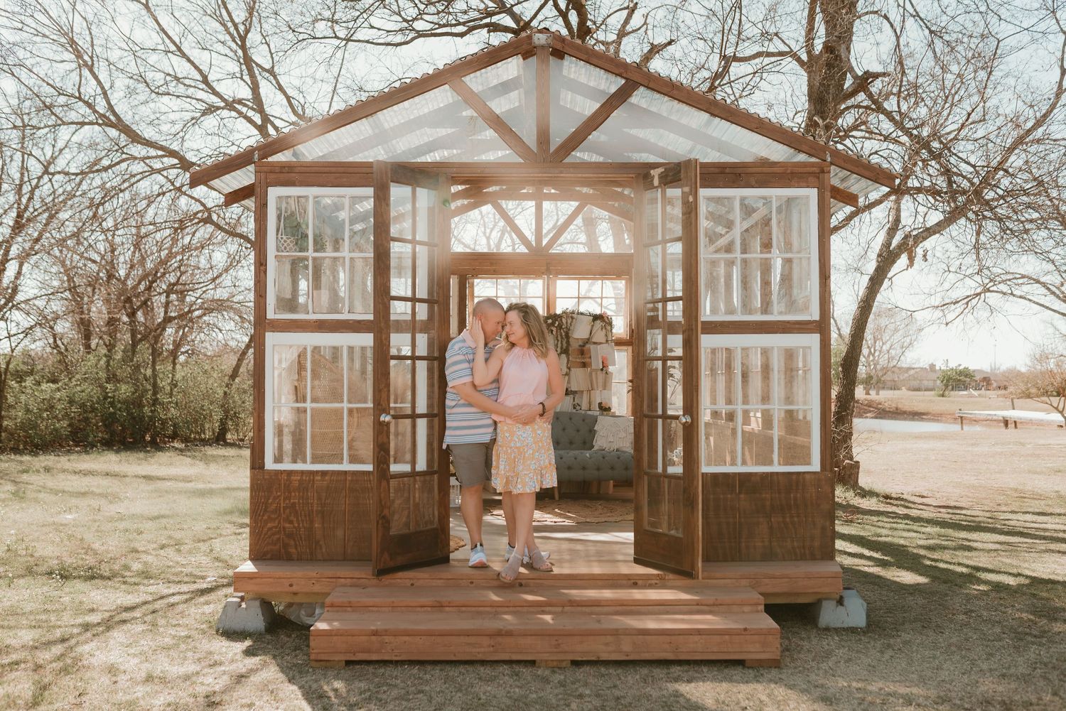 A couple embraces inside a charming rustic greenhouse with large windows and wooden frame outdoors.