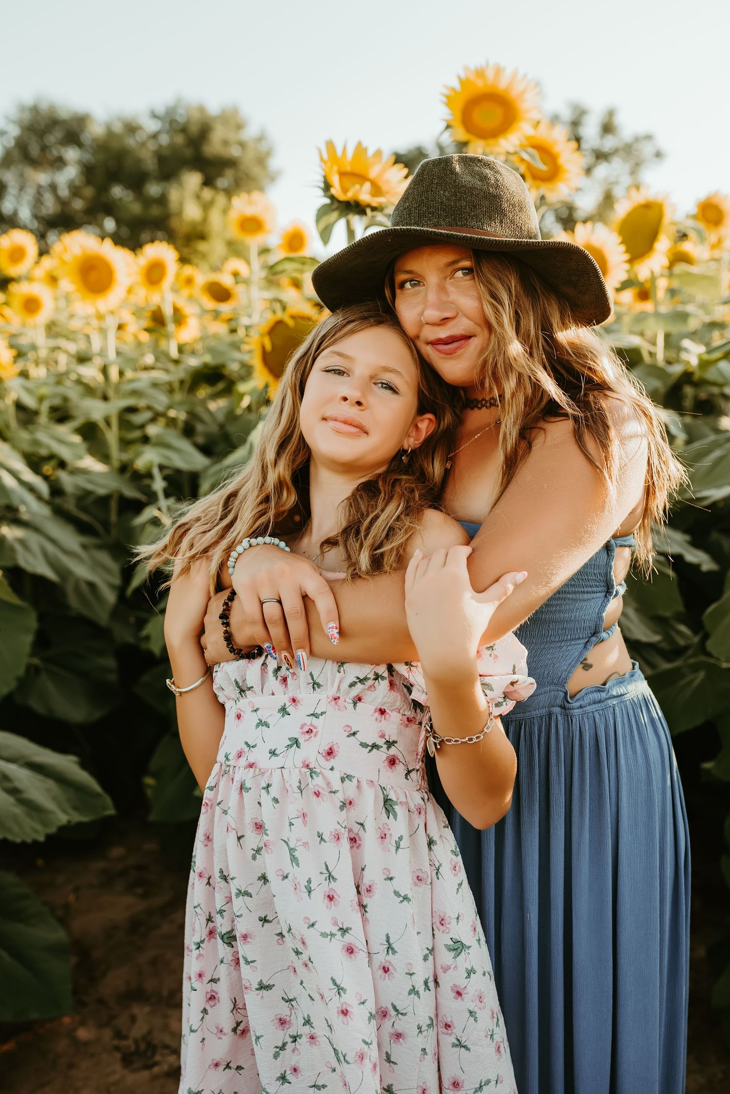 Woman in blue dress and hat hugs young girl in floral dress in a sunflower field.