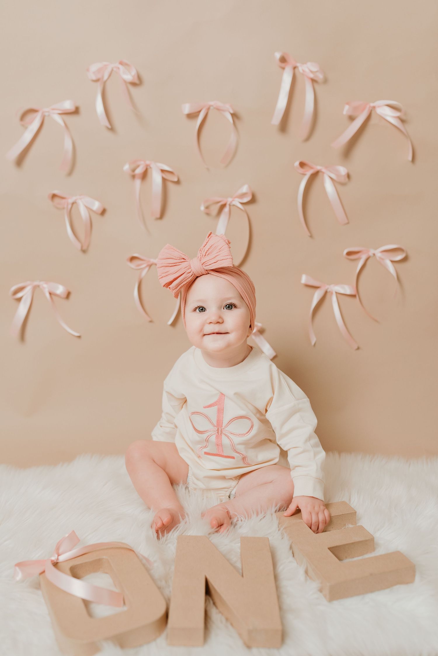 One year old baby girl in cream birthday outfit sitting with ONE letters and pink bows on beige backdrop.