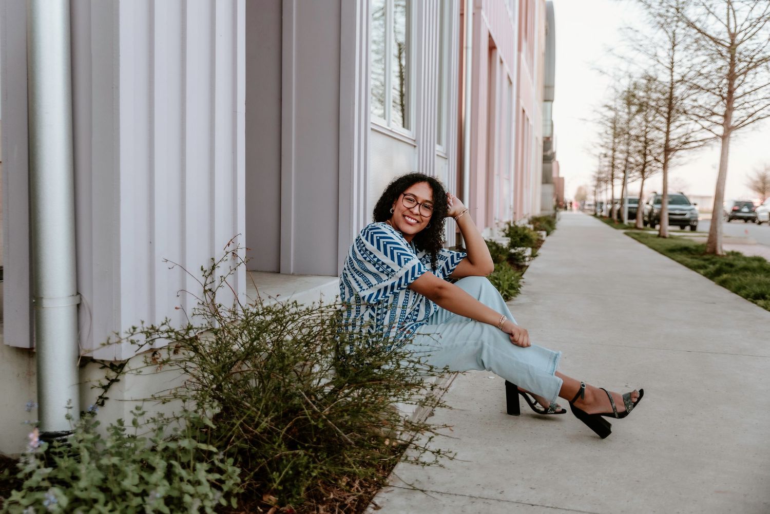 Woman in blue patterned top and jeans sitting on sidewalk curb beside modern colorful buildings.