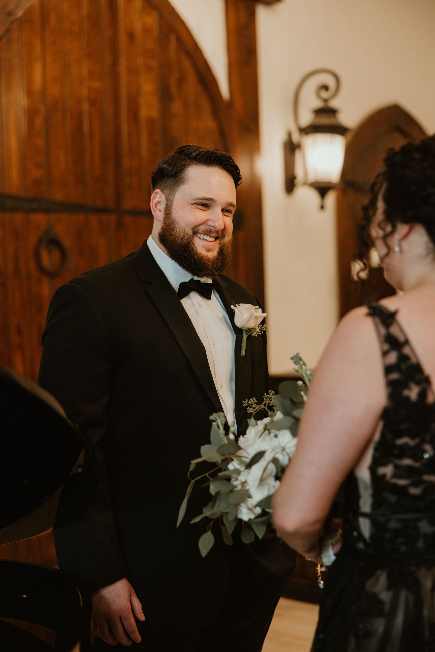 Smiling groom in tuxedo holds white boutonniere bouquet during first look with bride in black gown.