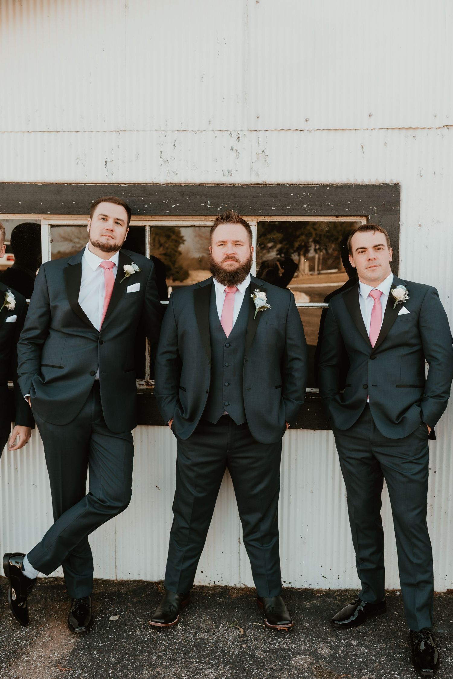 Three groomsmen in dark navy suits with pink ties posing together against a white barn wall.