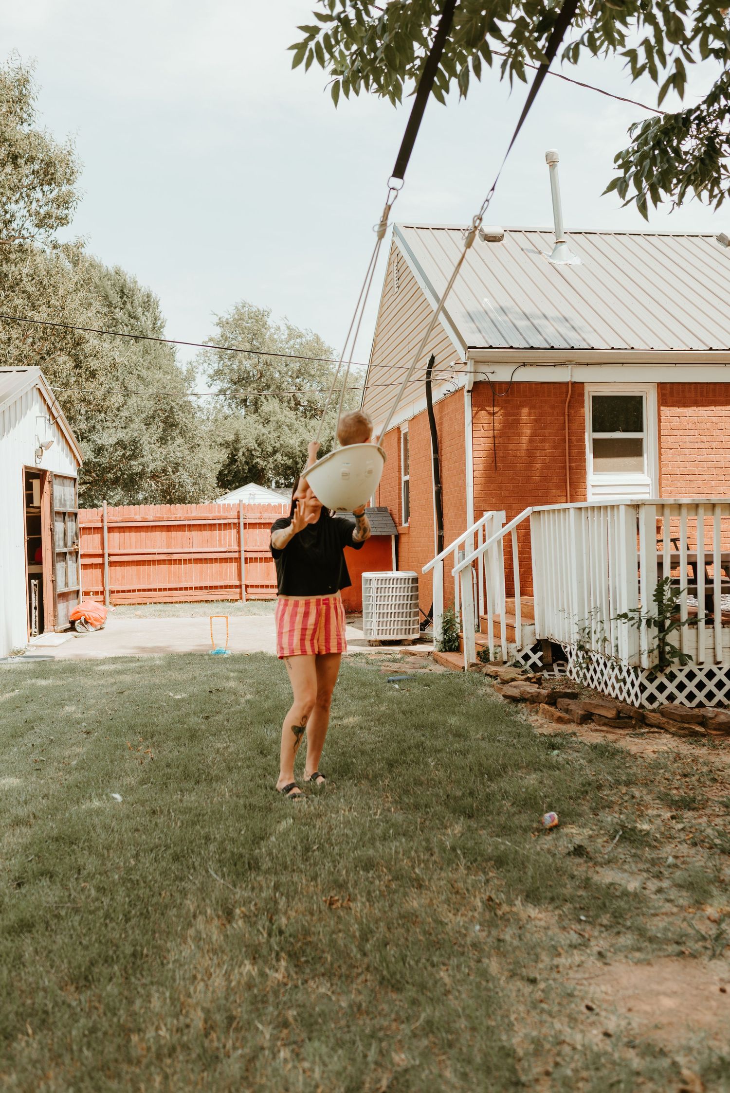 Woman in pink skirt carries a baby on her shoulders in a sunny backyard beside a brick house.