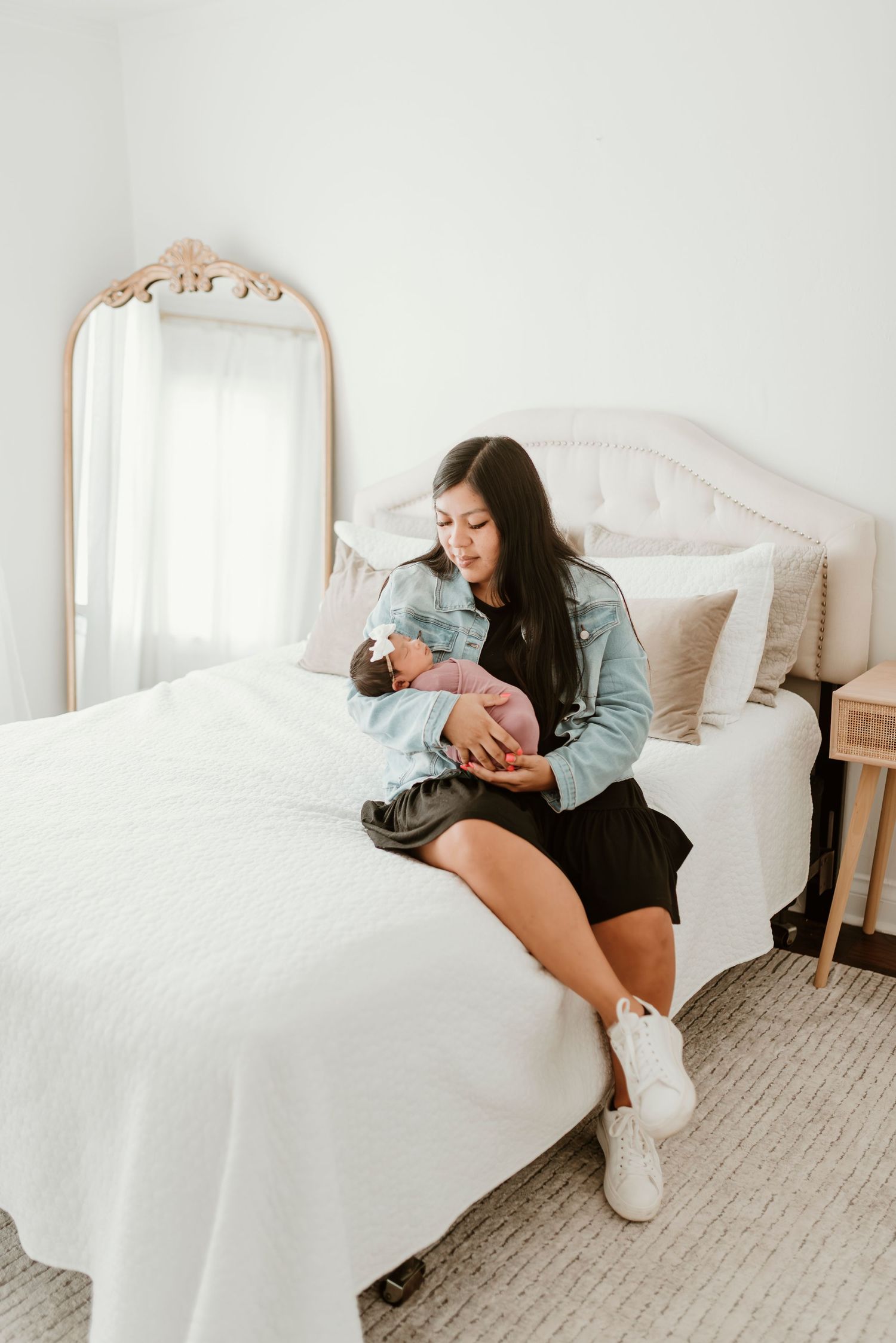 A dark-haired mother in a denim jacket sits on a white bed, tenderly cradling her sleeping newborn baby.