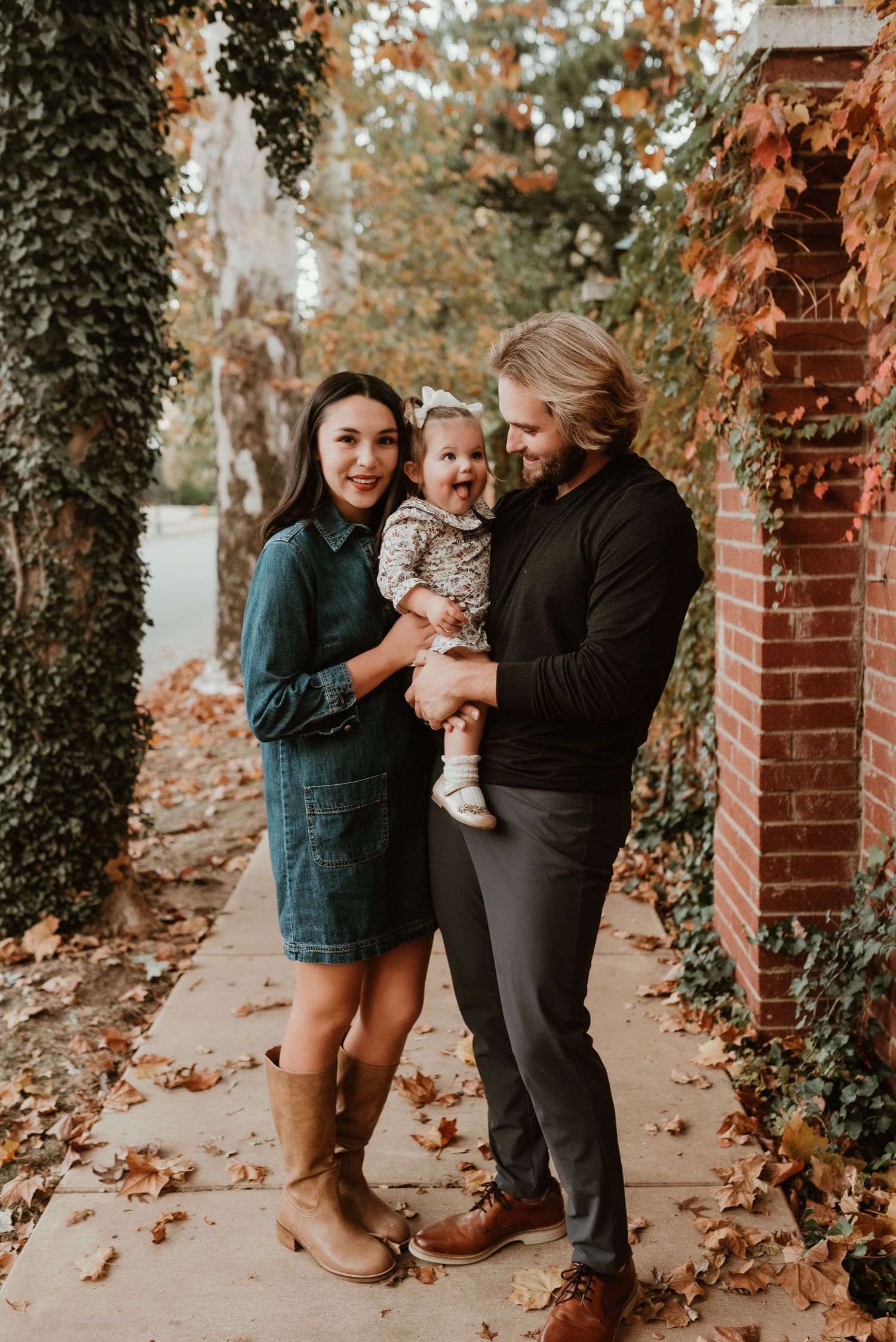 Smiling couple holds baby girl while standing on leaf-covered sidewalk in fall.