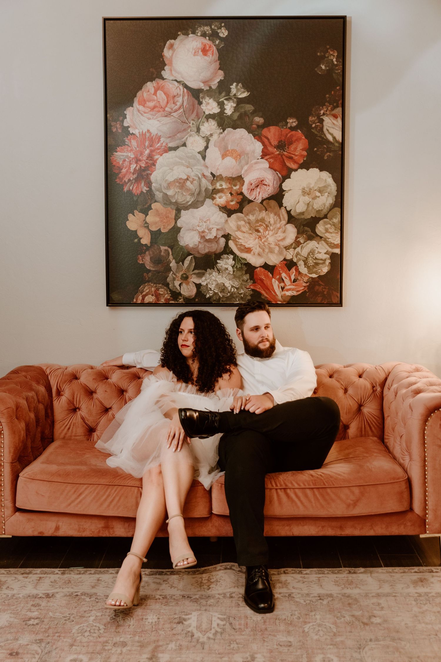 Engaged couple posing on a tufted velvet sofa beneath a large floral painting in a stylish interior.
