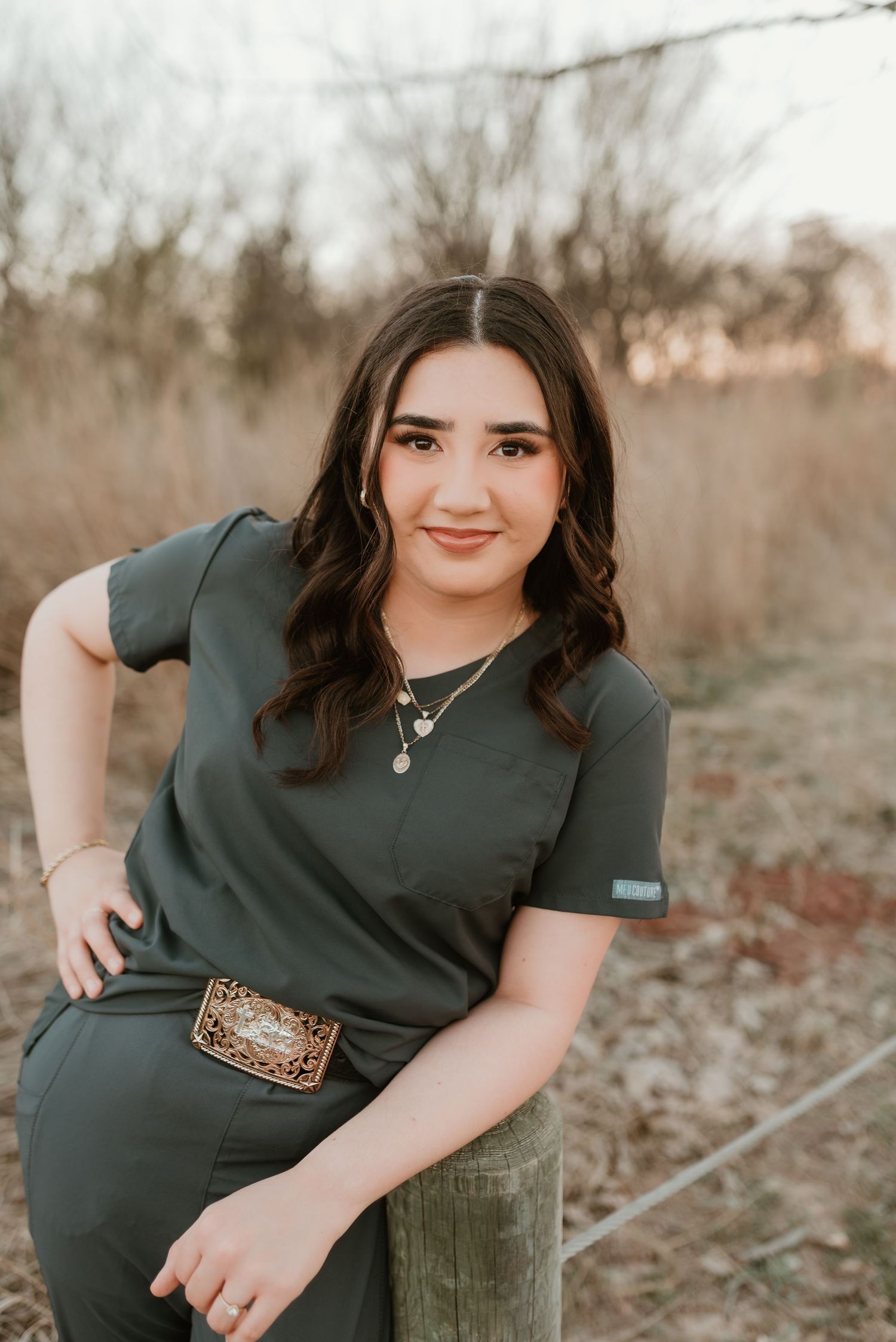 Brunette woman in olive green scrubs with leopard belt buckle sitting on post in natural setting.