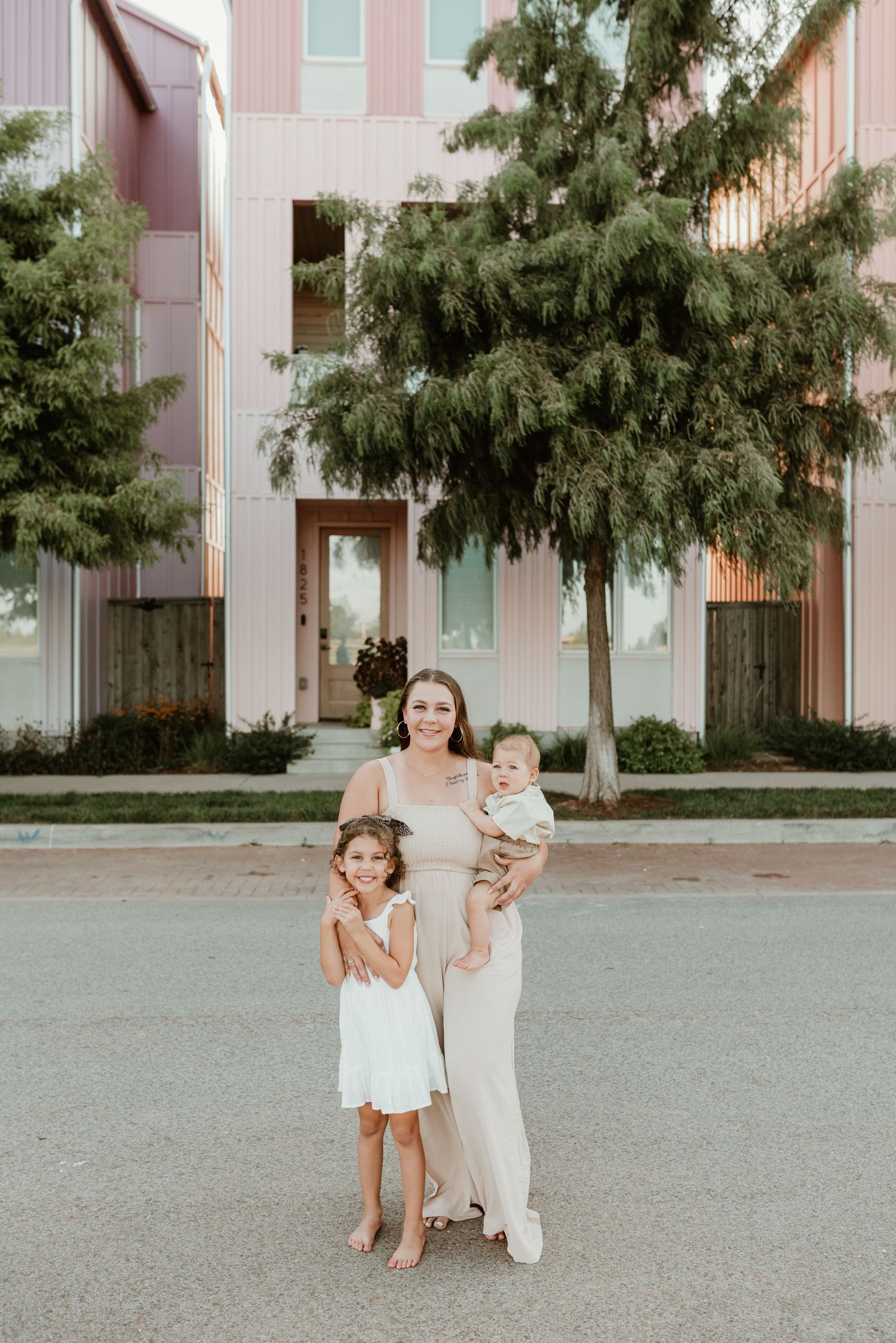 A smiling mother in a beige dress holds her baby while her young daughter stands beside her outdoors.