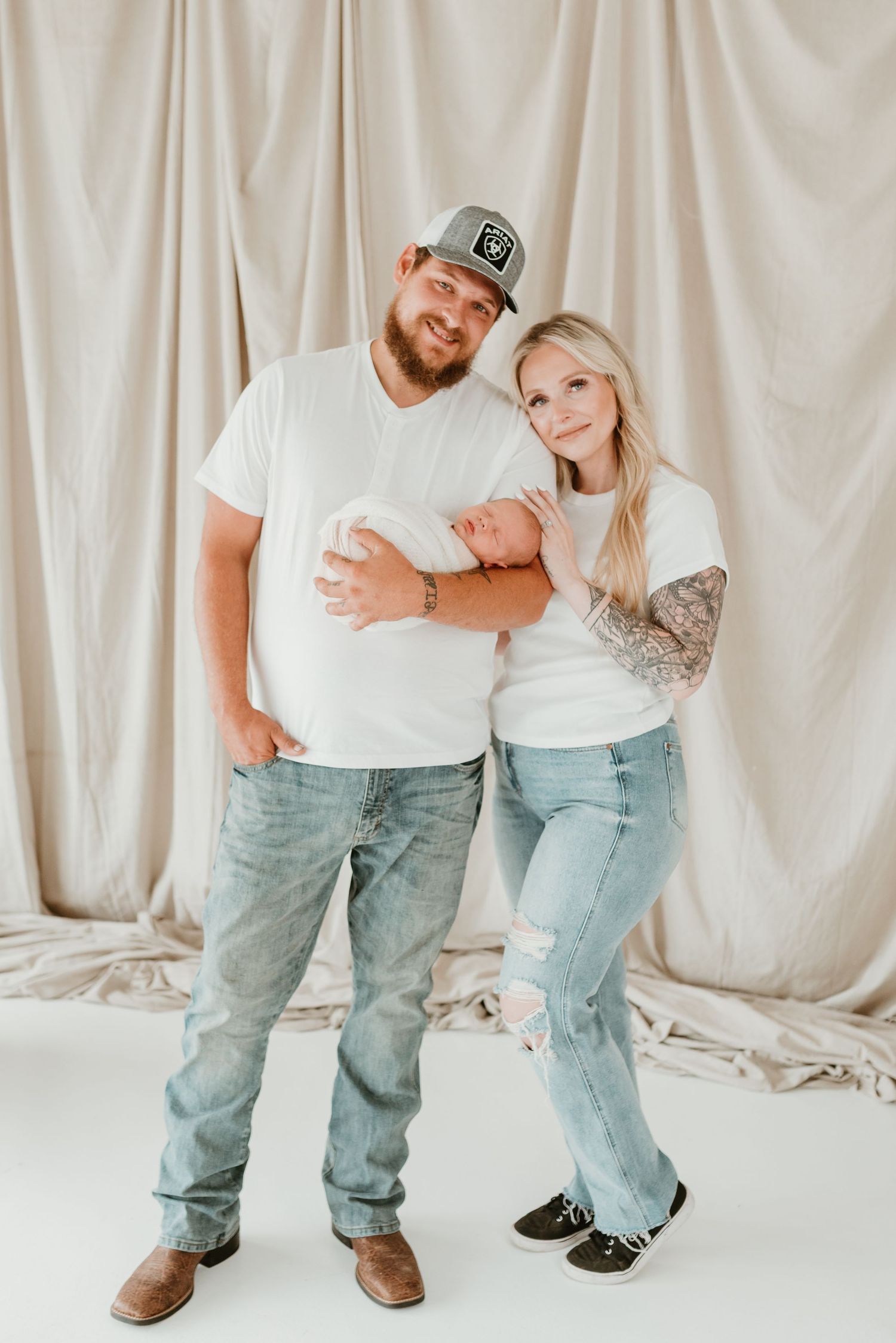 New parents in matching white shirts and jeans holding swaddled newborn in a bright studio.